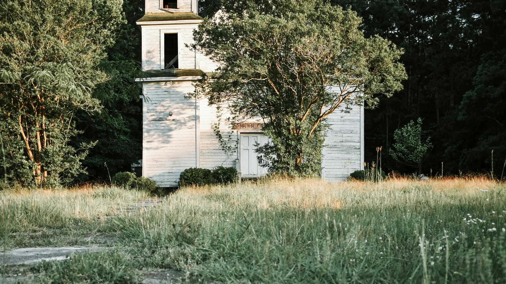 Old white wooden church with tower surrounded by tall grass and trees in rural setting at sunset.