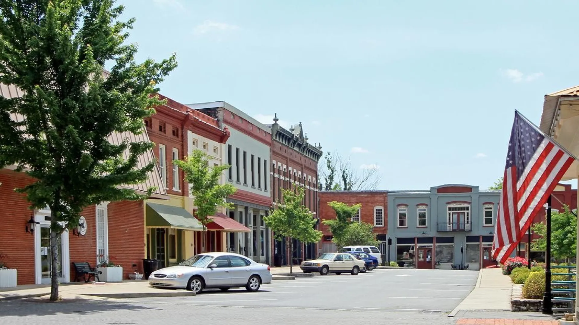 Small town main street with historic brick buildings, parked cars, trees, and American flags on a sunny day.