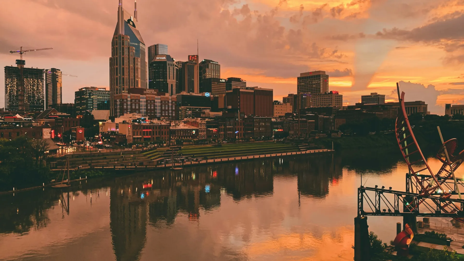 Nashville city skyline at sunset with buildings reflected in calm river waters under colorful sky.