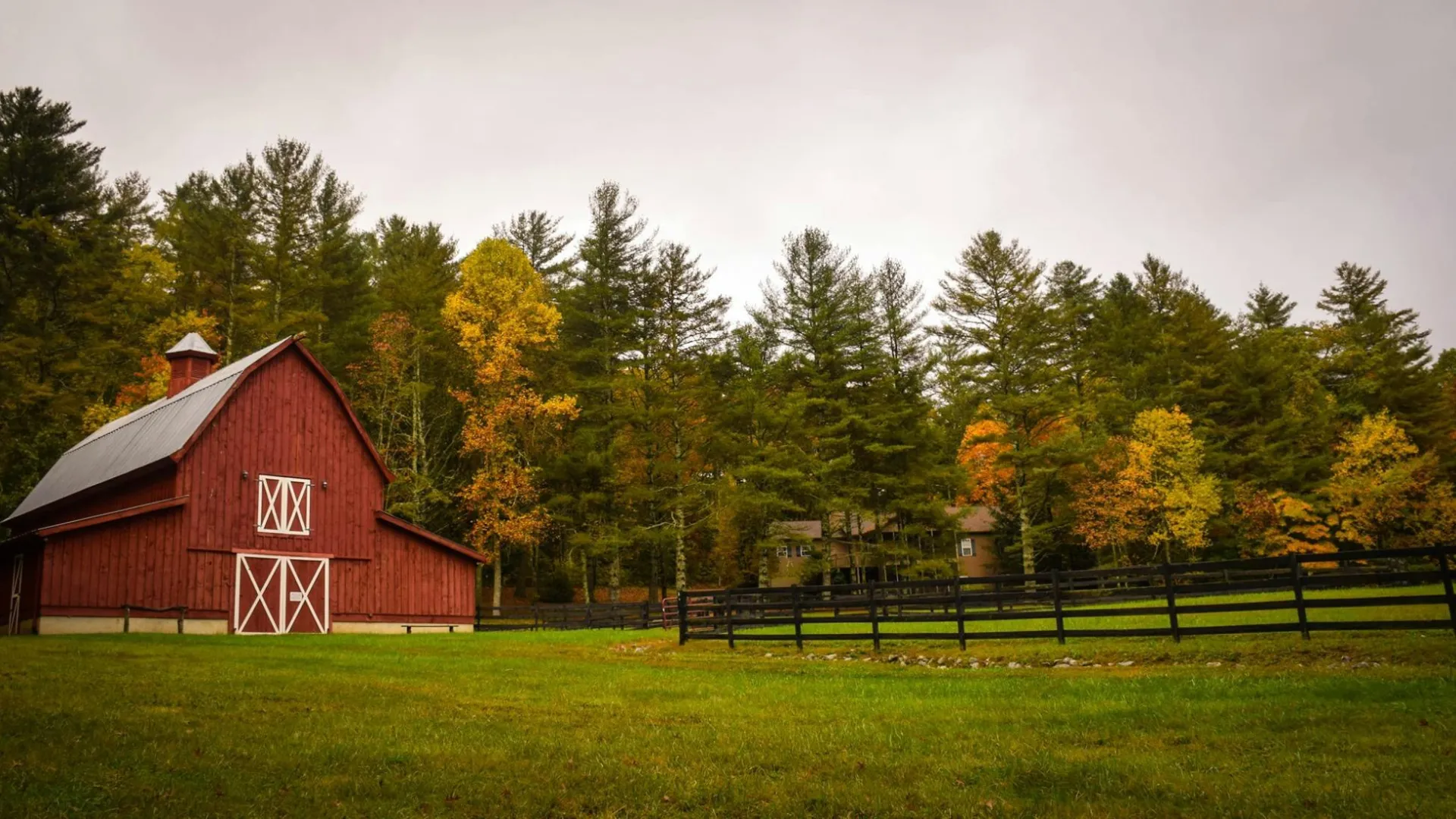 Red barn near wooden fence with green grass and autumn trees under cloudy sky in rural landscape