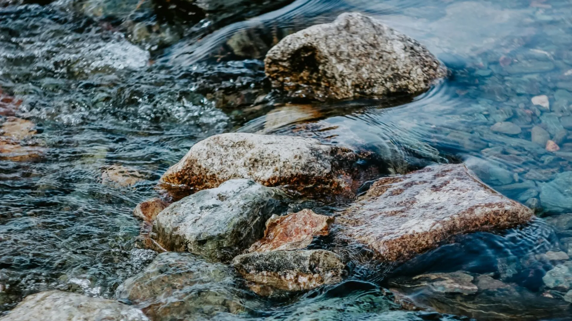 Clear water flowing over smooth multicolored river rocks in a shallow stream with natural light reflections