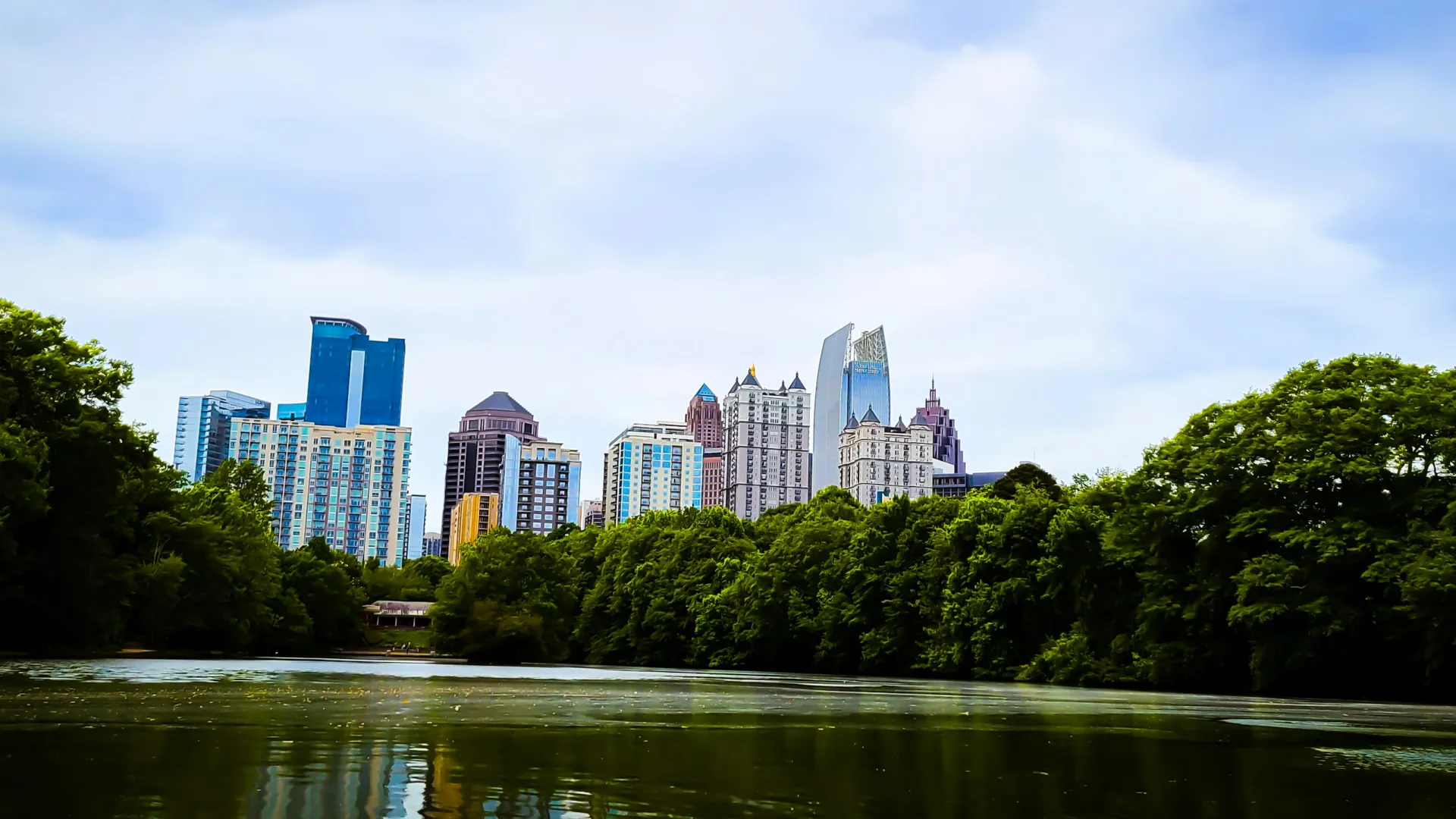 City skyline with modern buildings seen behind lush green trees by a calm river under a partly cloudy sky.