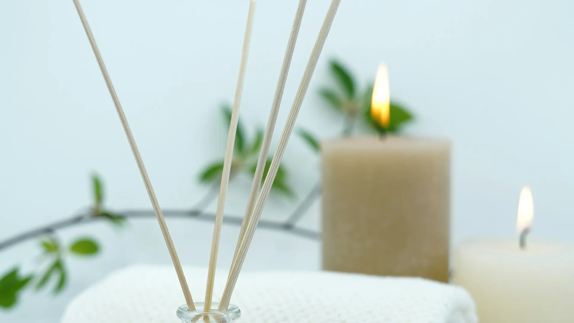 Aromatherapy diffuser with wooden sticks, rolled white towel, lit candles and green leaves for spa relaxation.