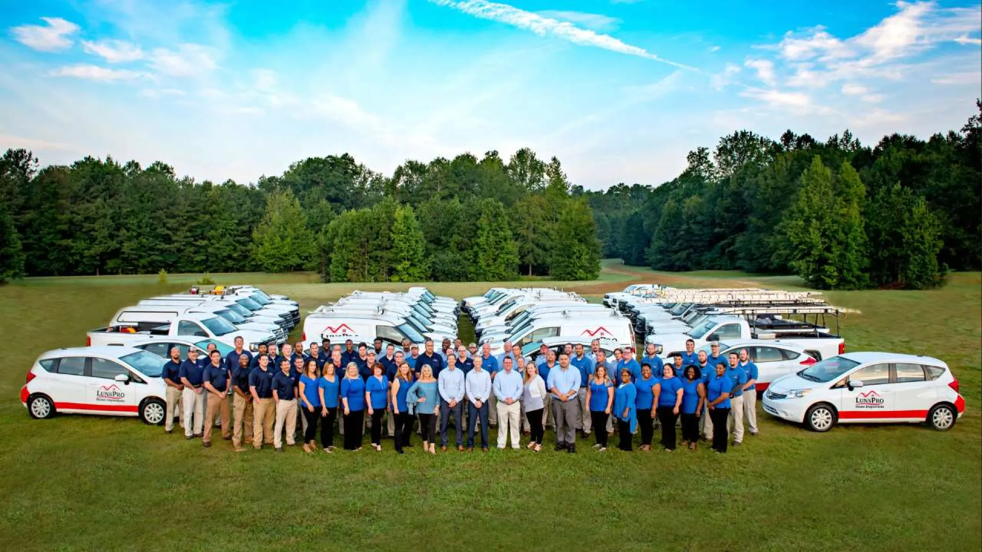 Large group of workers in blue uniforms standing in front of multiple white company vans on green field under blue sky.