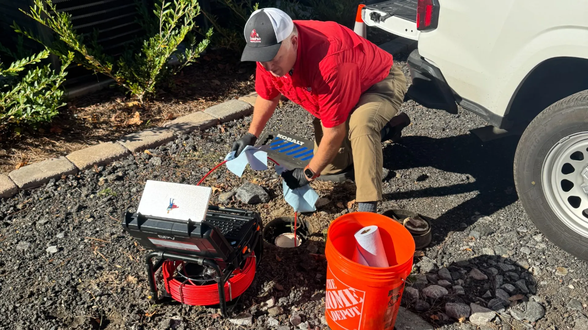 Technician in red shirt working with tools and equipment on rocky ground near white utility truck and orange bucket.