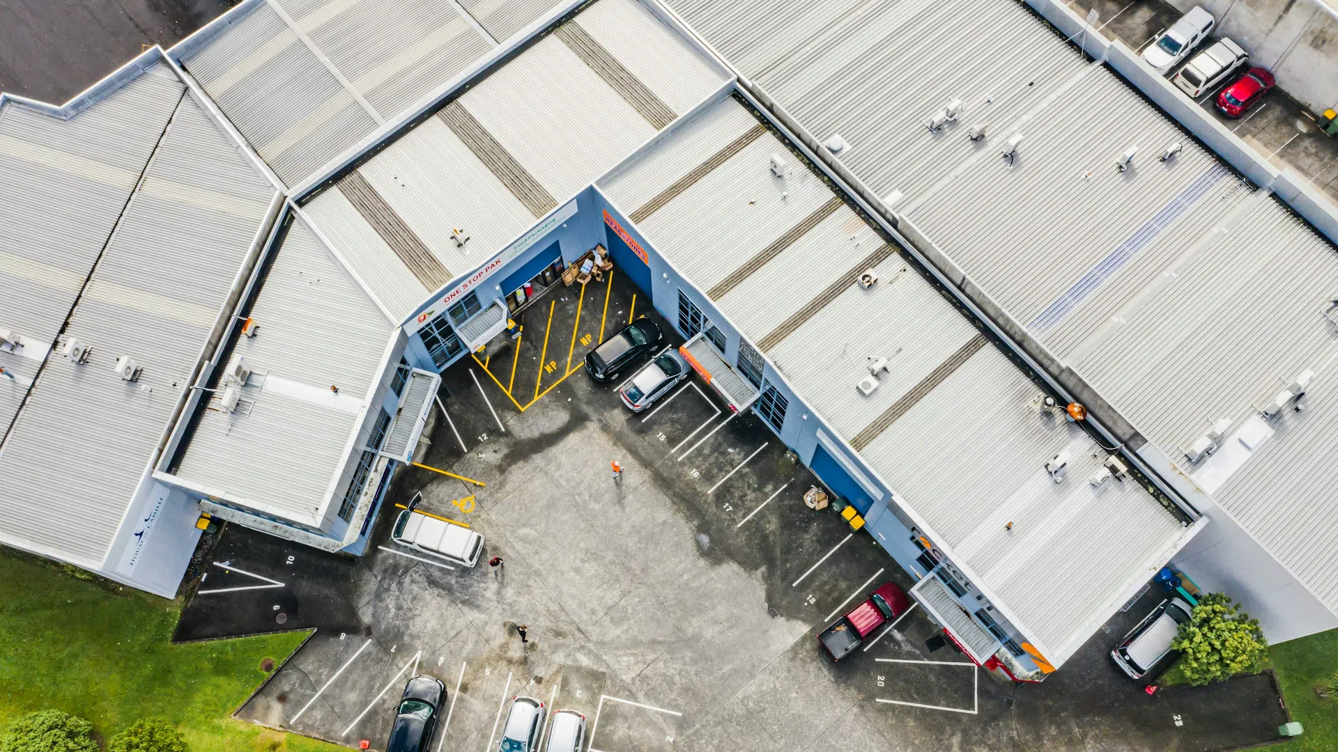 Aerial view of a commercial building with parking spaces and several parked cars surrounded by green grass and roads.