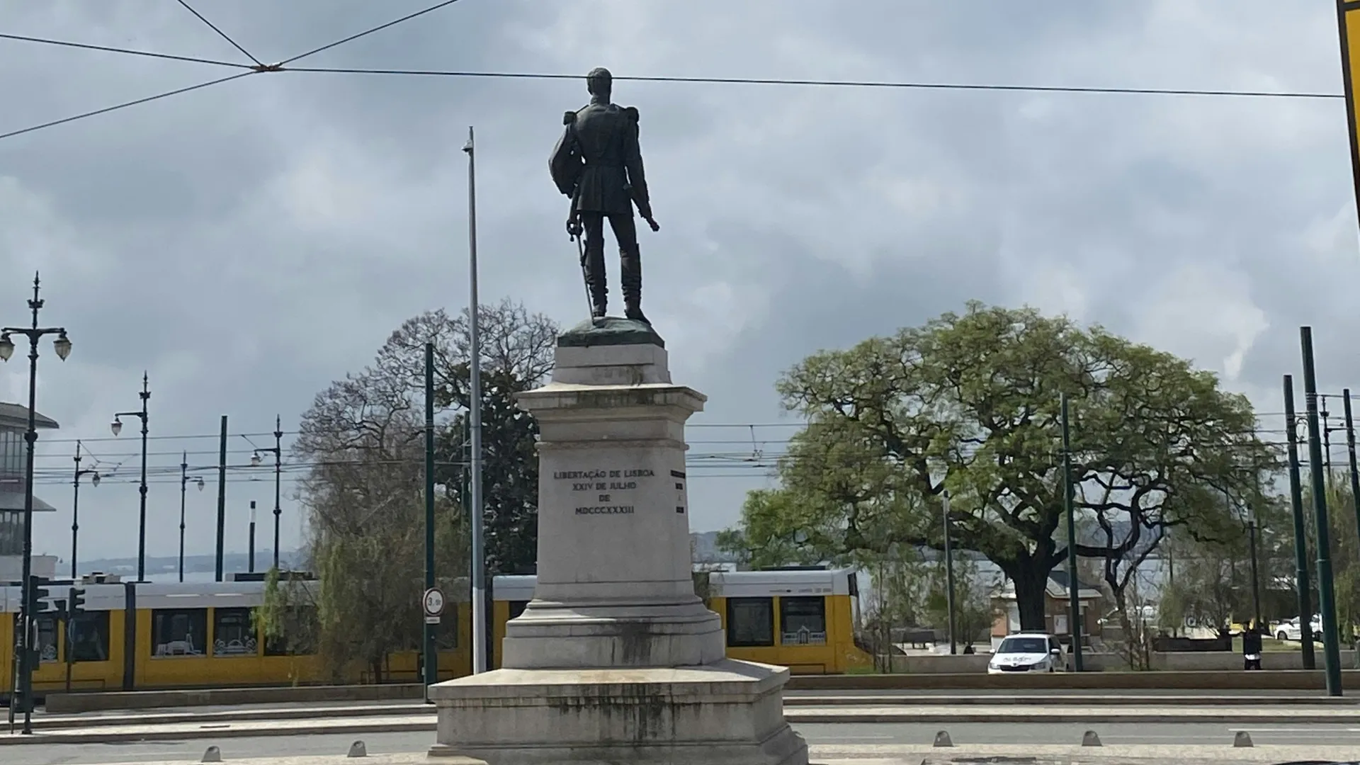 Statue on pedestal in open plaza with tram, cars, trees, and overhead wires under a cloudy sky