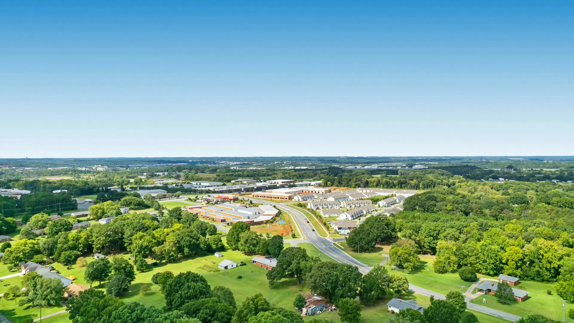 Aerial view of a suburban area with green trees, houses, curved roads, and clear blue sky on a sunny day.