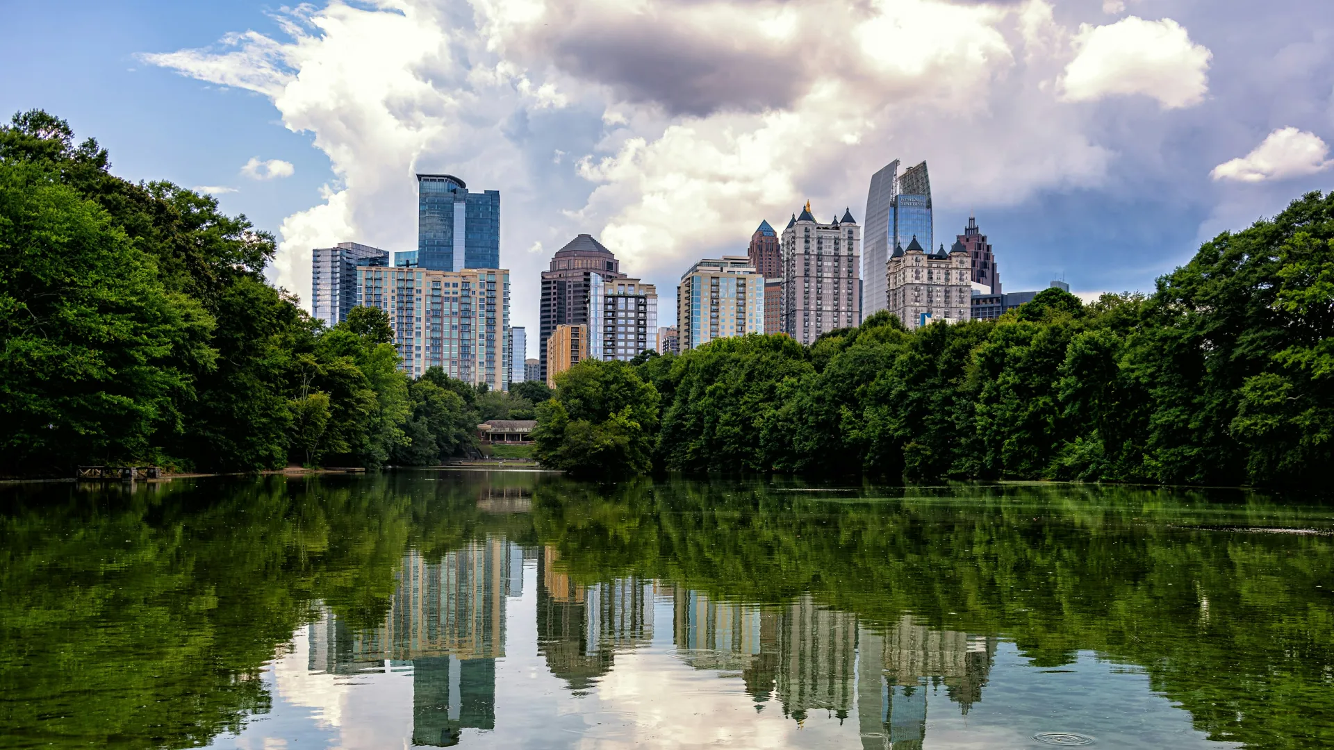 City skyline reflected in calm water with lush green trees under a cloudy sky during daytime.