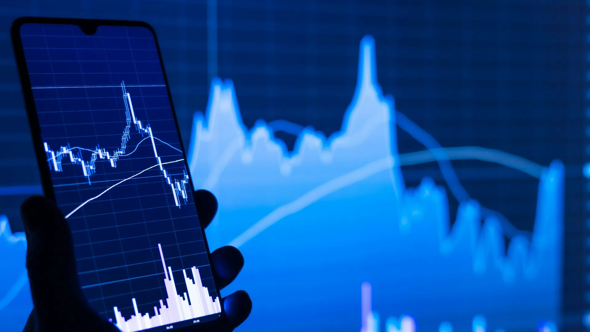 Hand holding smartphone displaying financial stock chart with graphs and trading data on blue background.