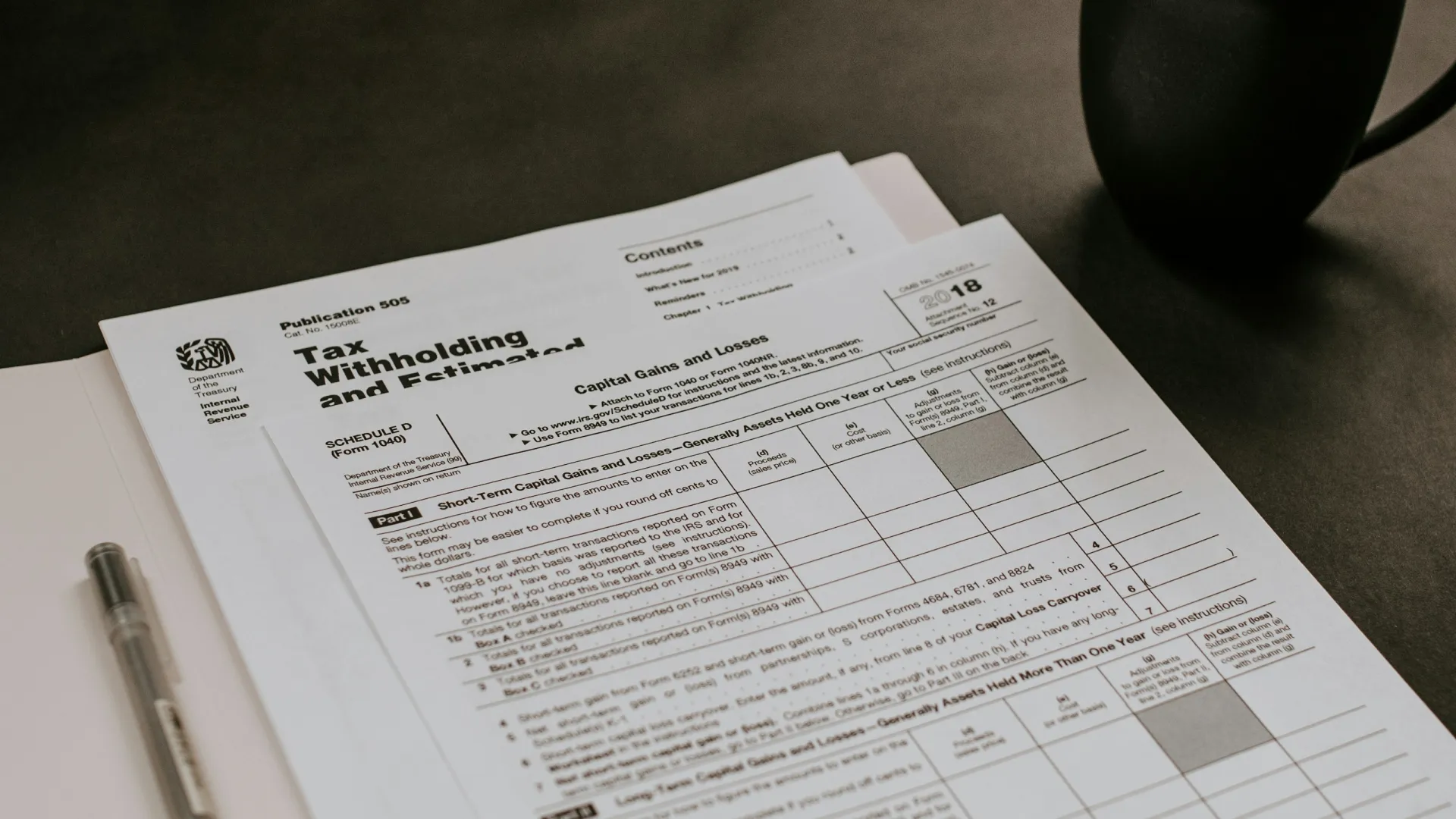 Tax withholding and estimate forms beside a pen and black coffee cup on a dark surface.