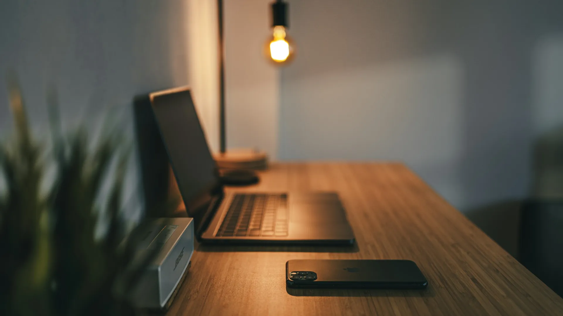 Modern workspace with a wooden desk, laptop, smartphone, desk lamp, and blurred plant in warm lighting.