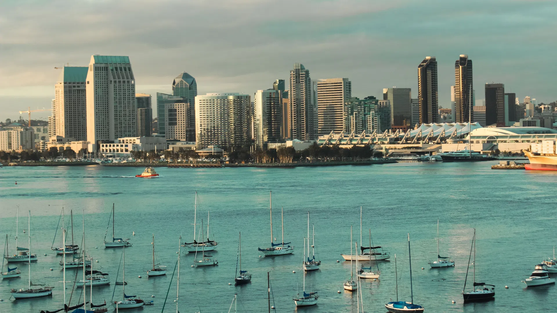 San Diego skyline with boats anchored in calm bay waters under a cloudy sky at golden hour.