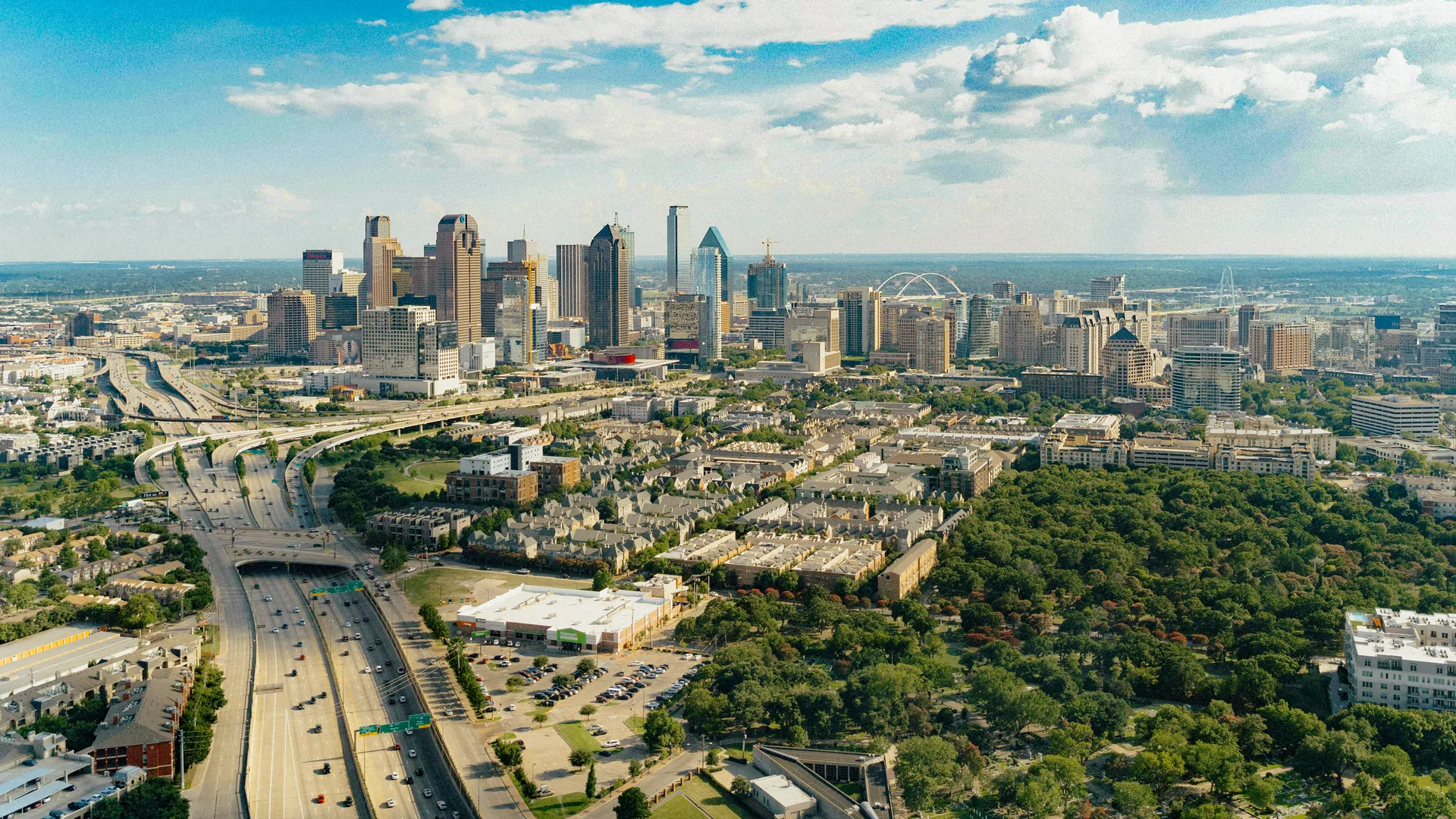 Aerial view of downtown Dallas skyline with highways, buildings, and a large urban park under a partly cloudy sky.