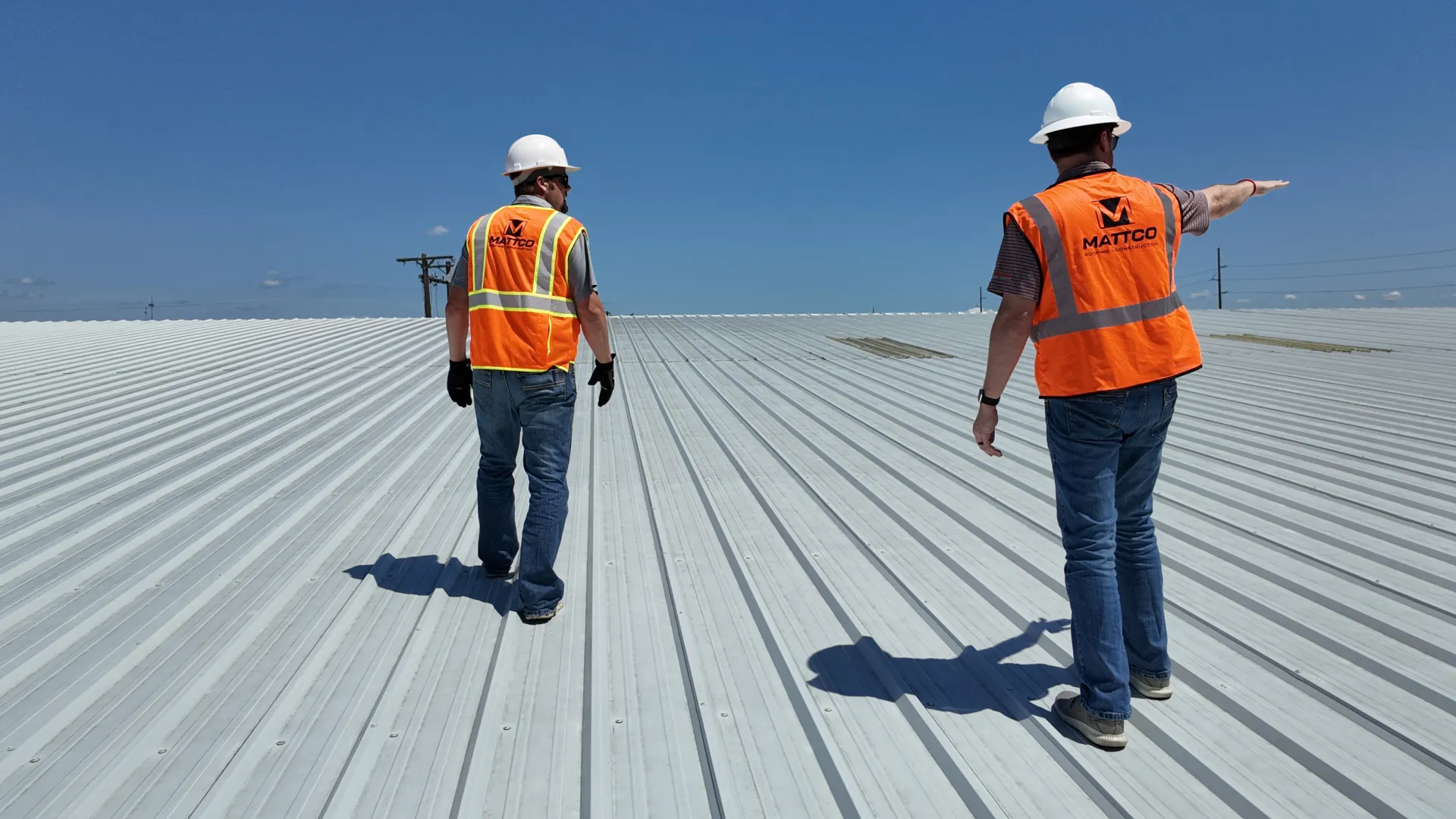 Two Mattco Roofing and Construction technicians in orange safety vests walking across a large commercial metal roof during a site inspection in Texas. One technician wearing a white hard hat is pointing across the roof surface while assessing the condition of the standing seam metal roofing system under a clear blue sky.
