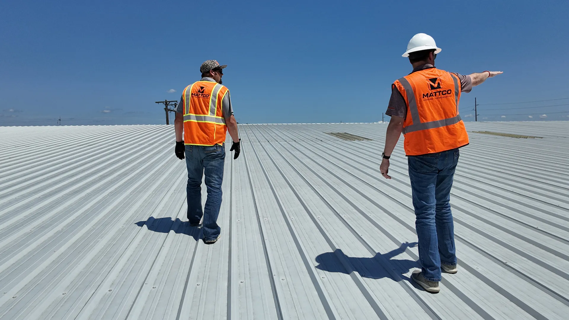 Two Mattco Roofing and Construction technicians in orange safety vests walking across a large commercial metal roof during a site inspection in Texas. One technician wearing a white hard hat is pointing across the roof surface while assessing the condition of the standing seam metal roofing system under a clear blue sky.