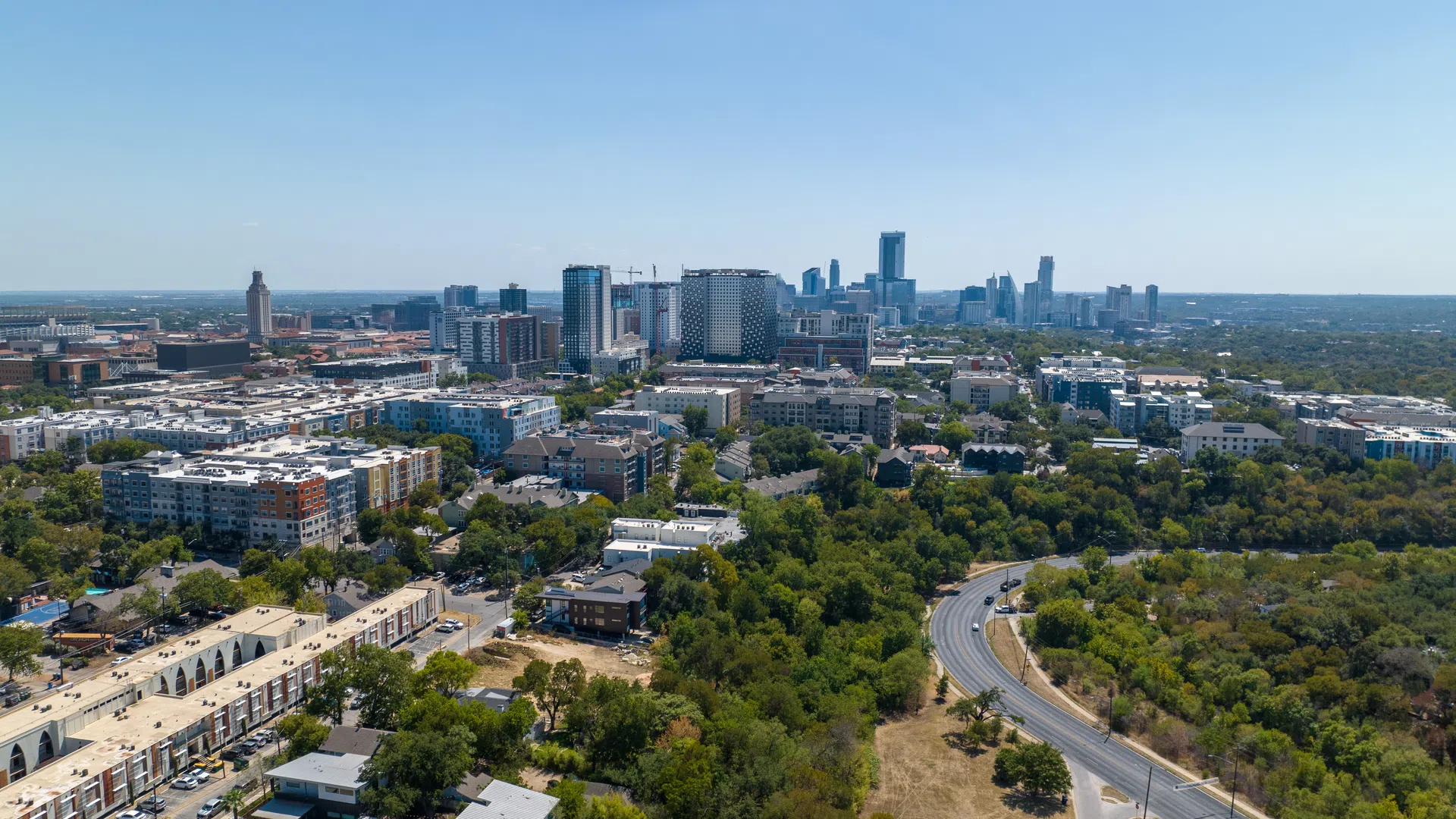 Aerial view of a city skyline with a mix of buildings and green spaces under a clear blue sky.