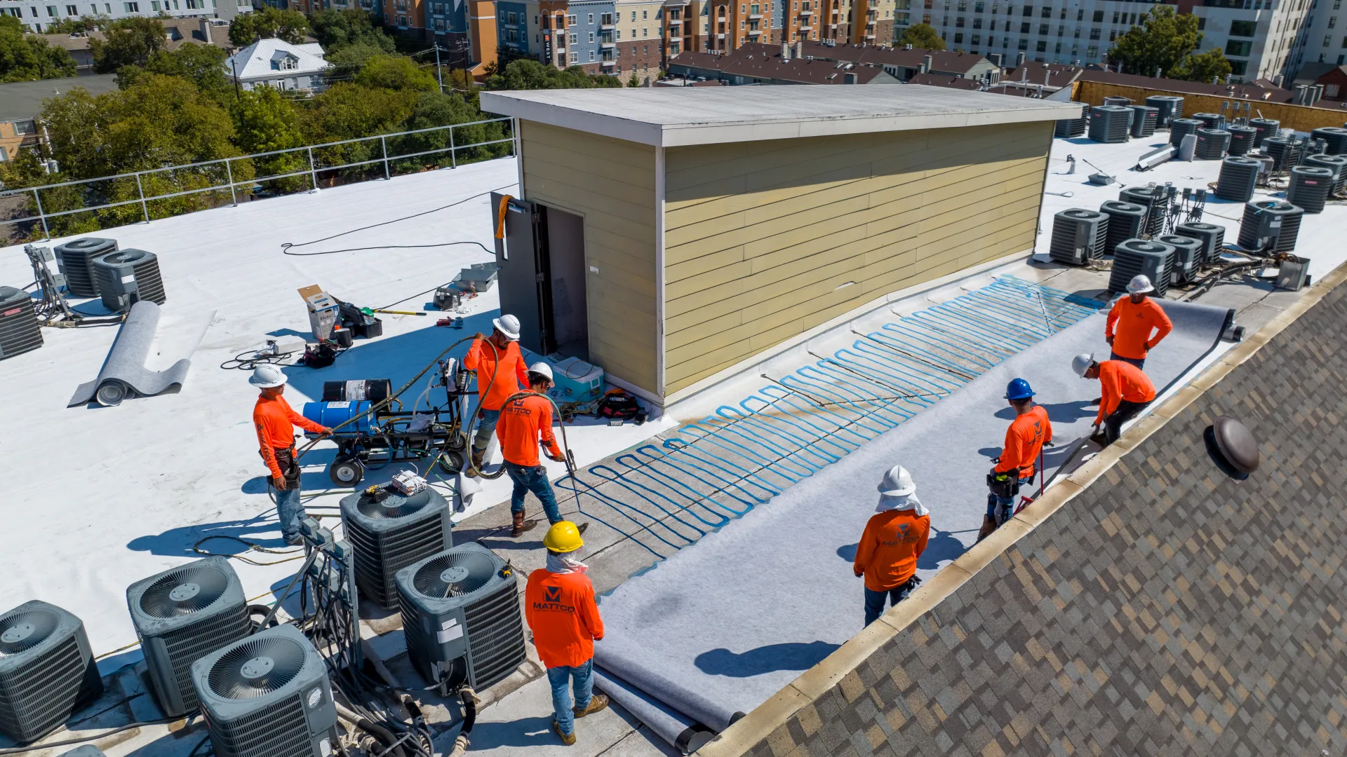 Construction workers in orange shirts installing heating elements on a white flat rooftop with HVAC units.