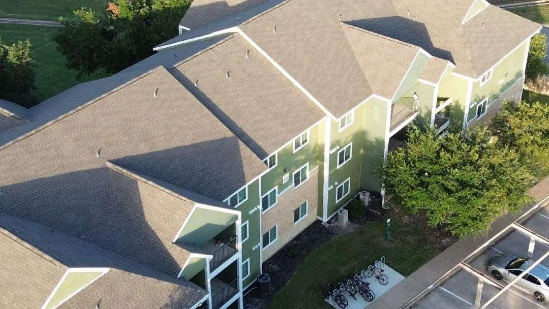 Aerial view of a green apartment complex surrounded by trees with adjacent parking and bike racks visible.