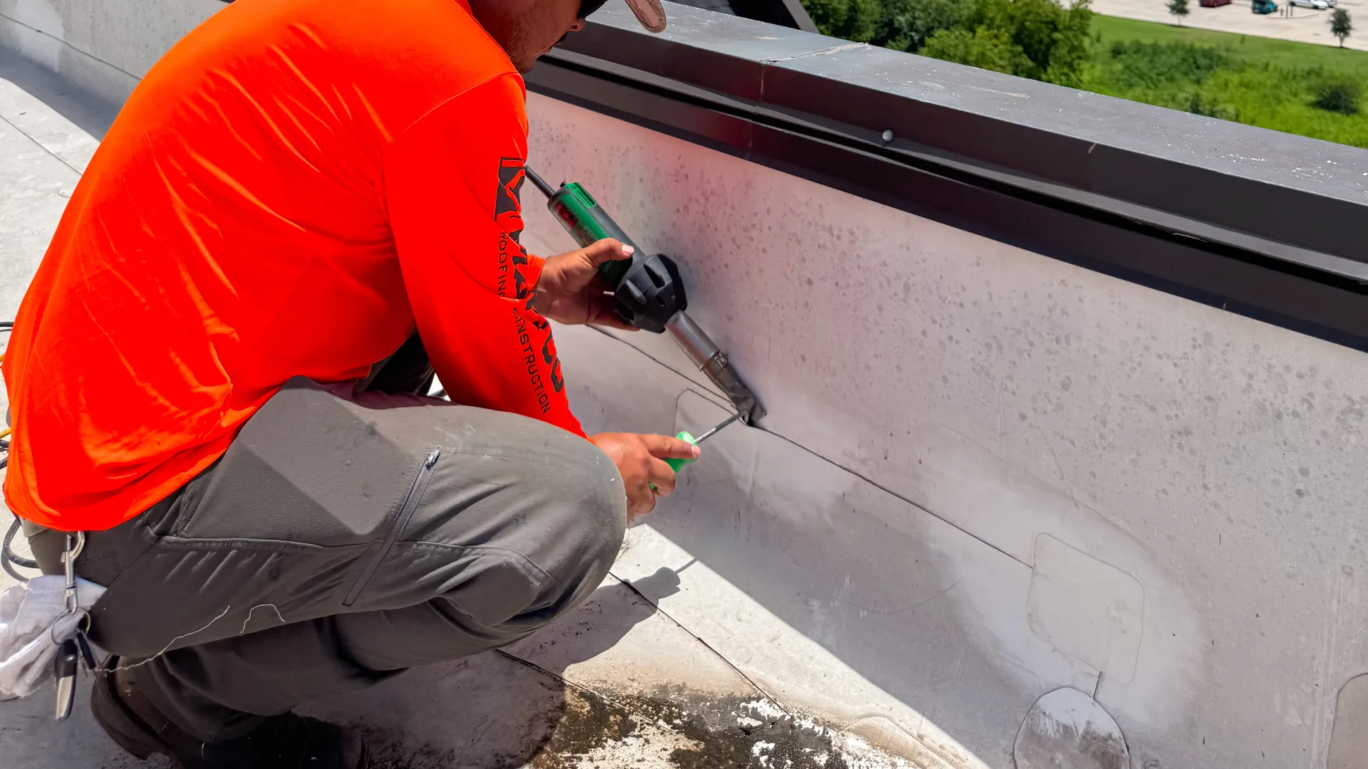 Worker in an orange shirt using a heat gun to seal a rooftop edge with city buildings in the background under blue sky.