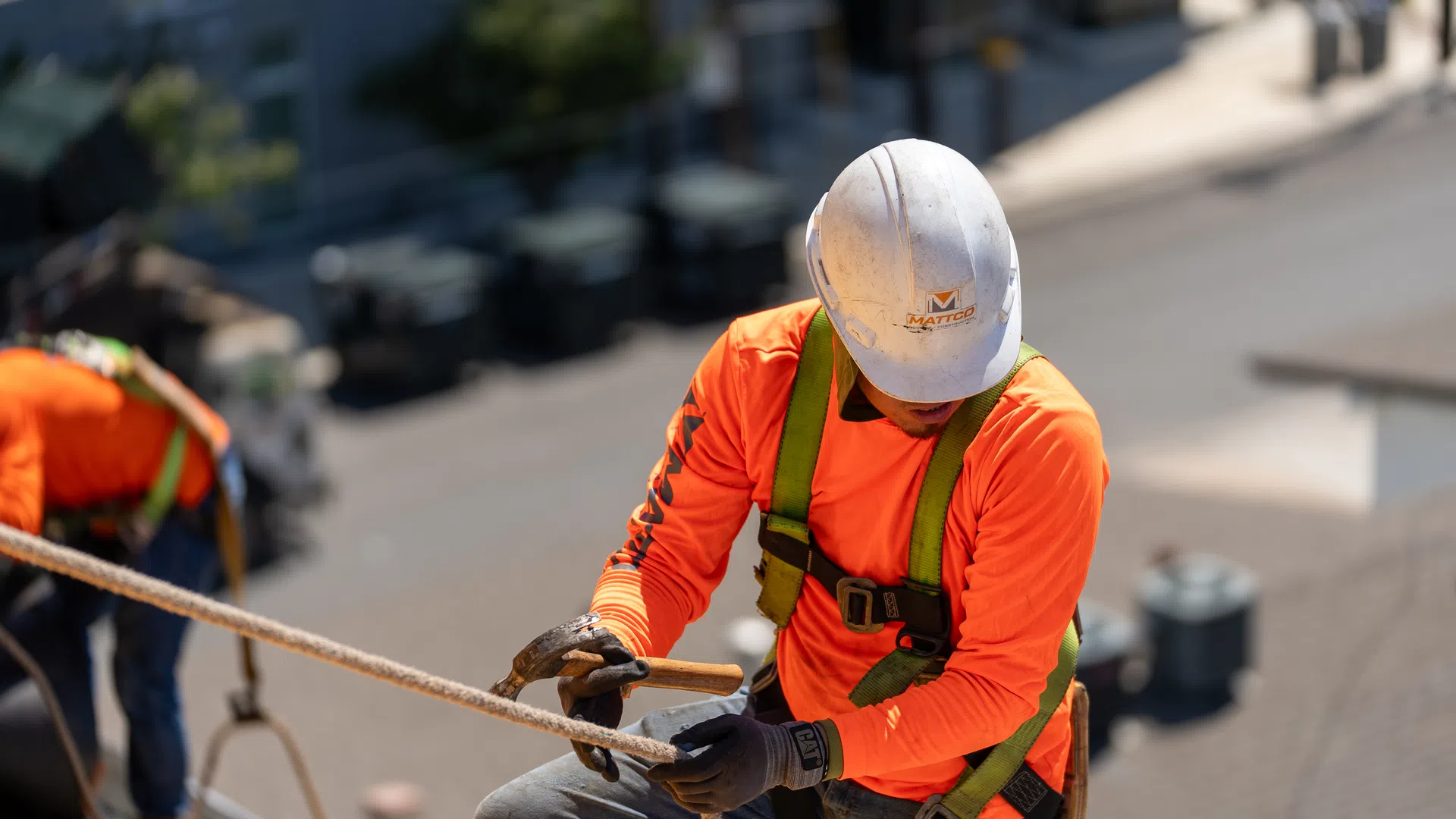 Construction worker in safety gear securing rope on building site wearing orange shirt and white hard hat.