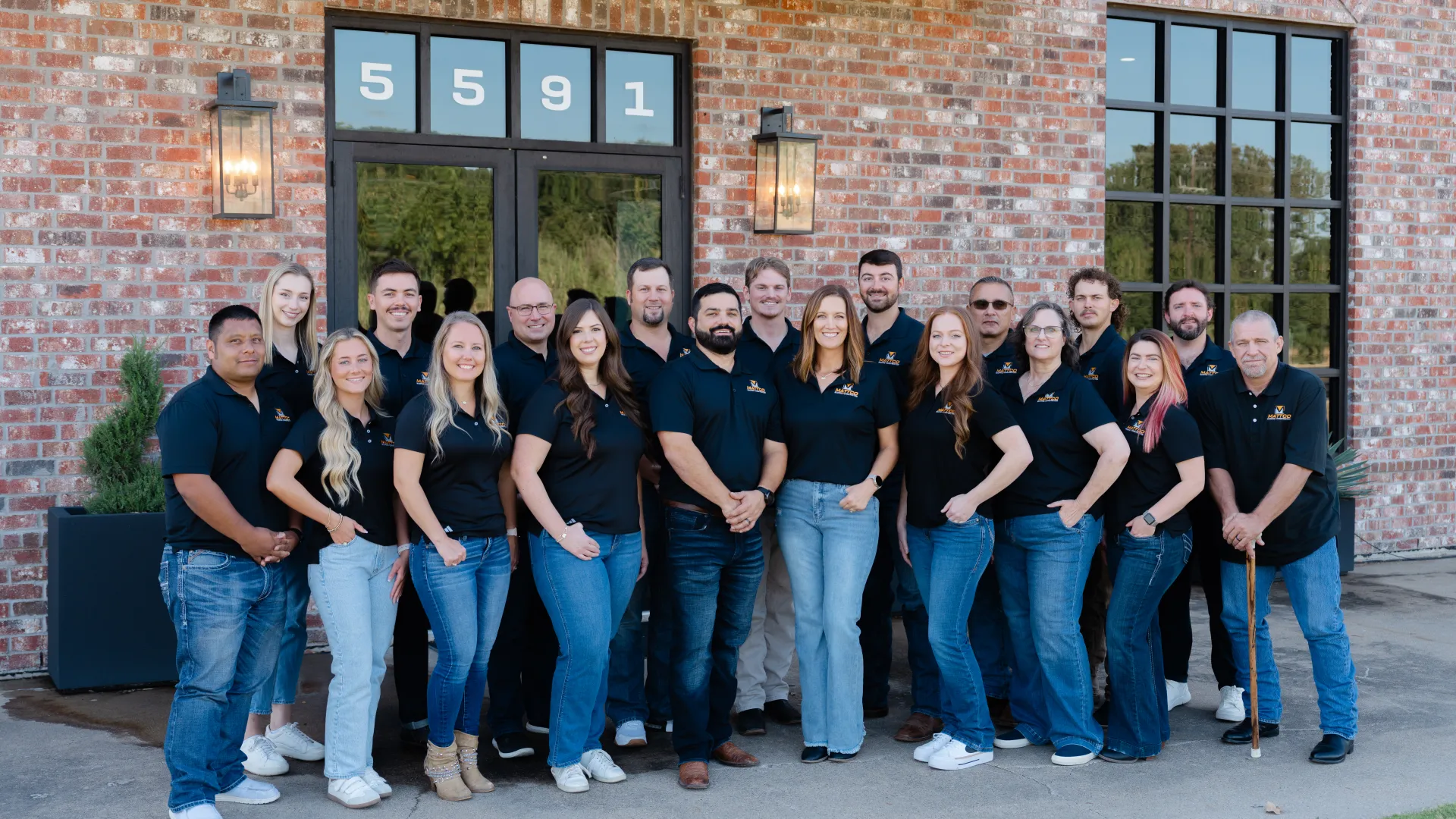 Group portrait of 19 diverse employees in black shirts standing outside a brick building with door number 5591.