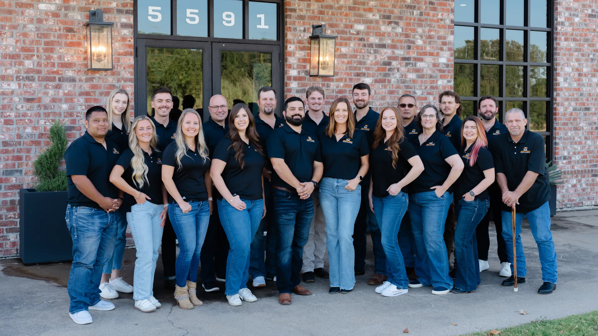 Group portrait of 19 diverse employees in black shirts standing outside a brick building with door number 5591.