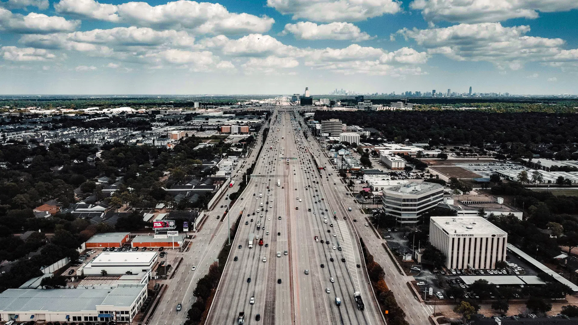 Aerial view of a busy highway stretching through a city with blue sky and scattered clouds above.