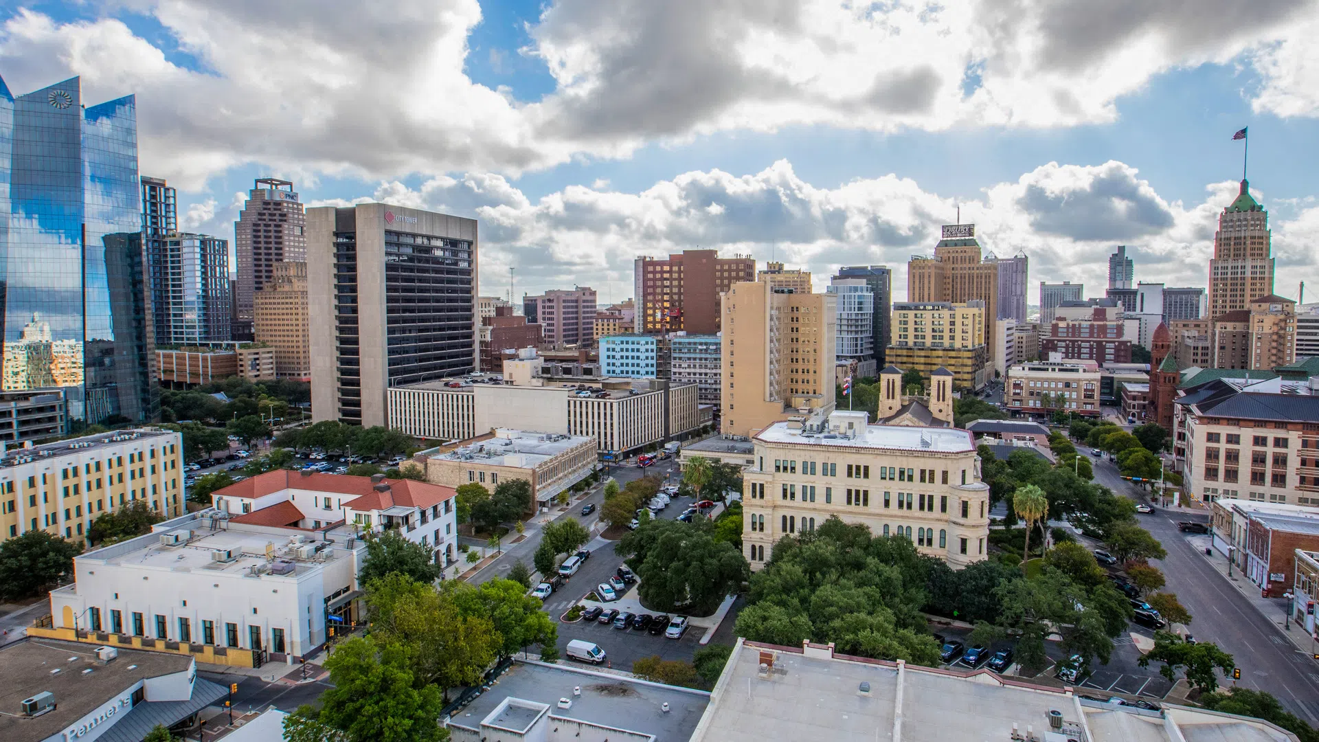 Aerial view of downtown San Antonio featuring modern skyscrapers, historic buildings, and a partly cloudy sky.