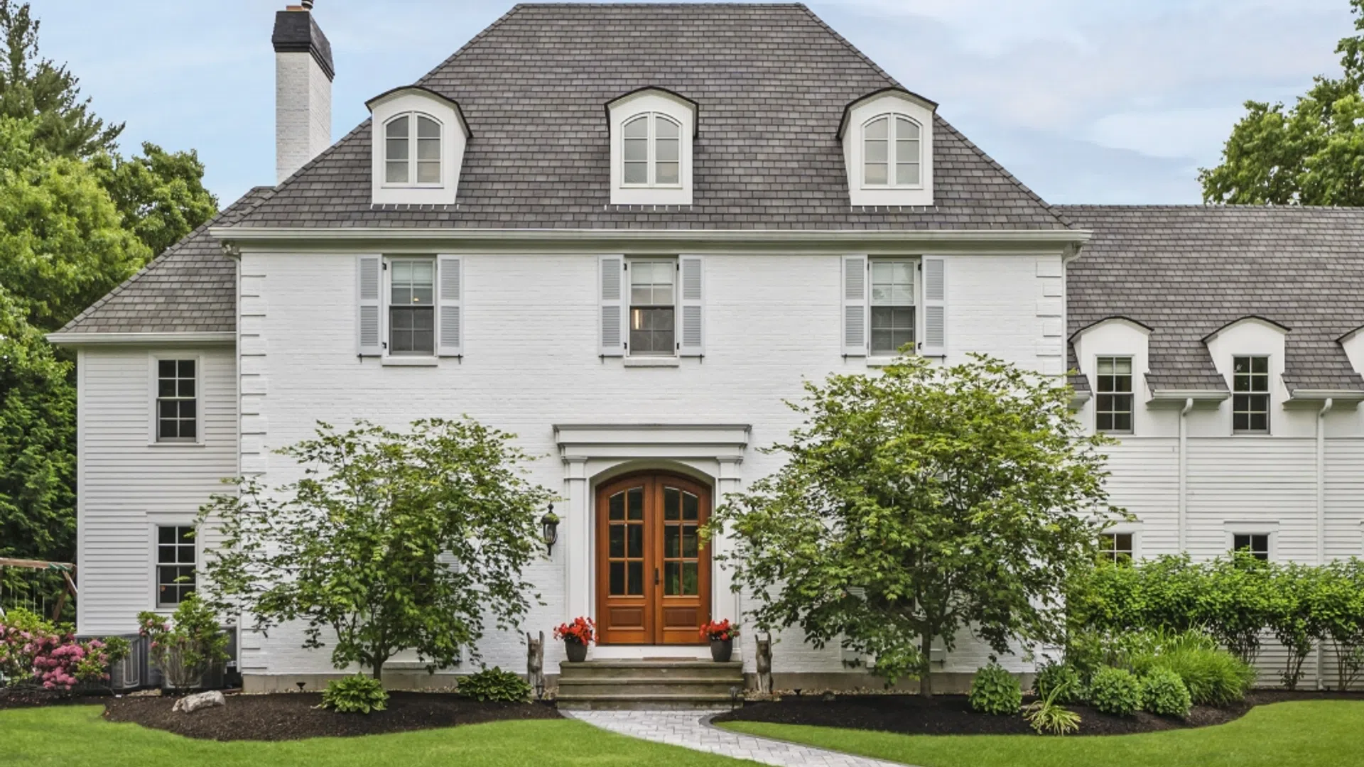 White two-story house with gray roof, wooden front door, landscaped lawn, and green trees under blue sky.