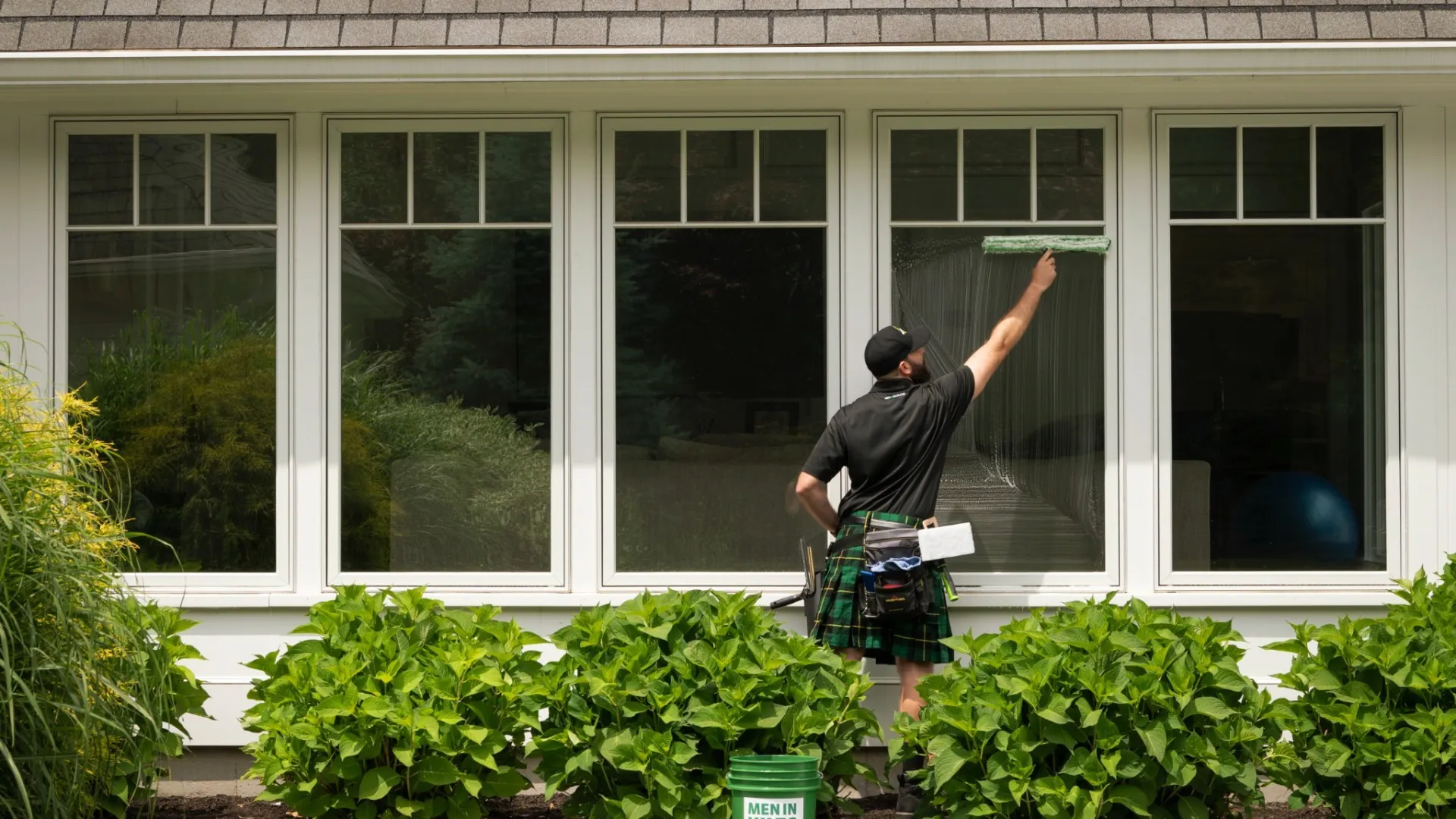 Man in a kilt cleaning large house windows with a squeegee outside green bushes and lawn