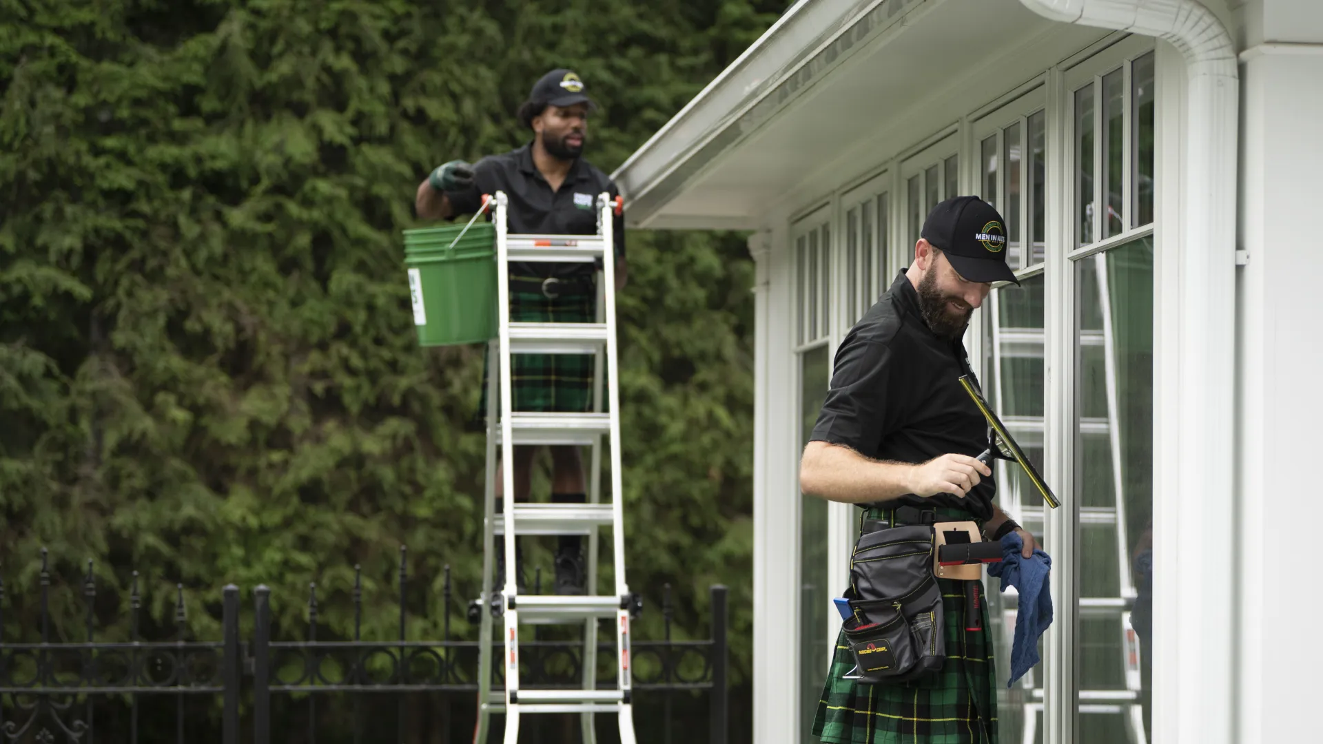 Two workers wearing kilts cleaning large windows outside a house, one on ladder, one wiping glass with squeegee.