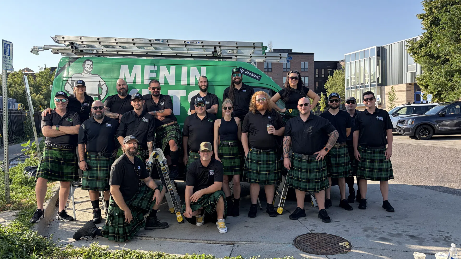 Group of workers wearing black shirts and green kilts posing in front of a green Men In Kilts service van outdoors.