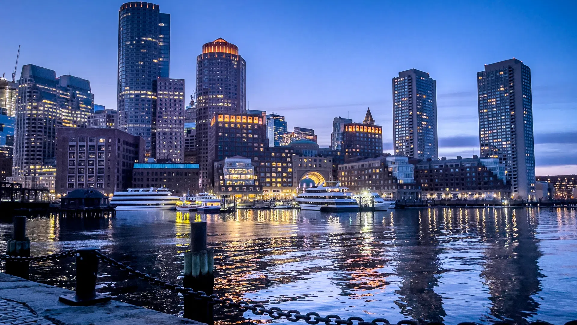 Boston skyline at dusk with illuminated skyscrapers reflecting on calm harbor water and dock in foreground