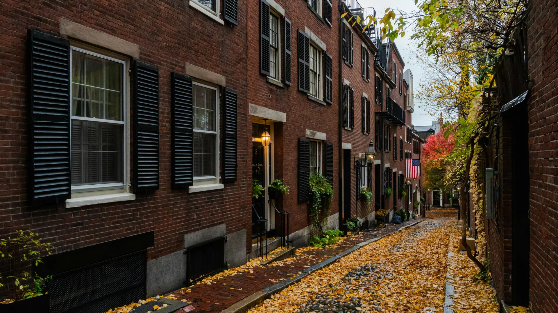 Cobblestone street lined with brick buildings and black shutters covered in autumn leaves under a cloudy sky.