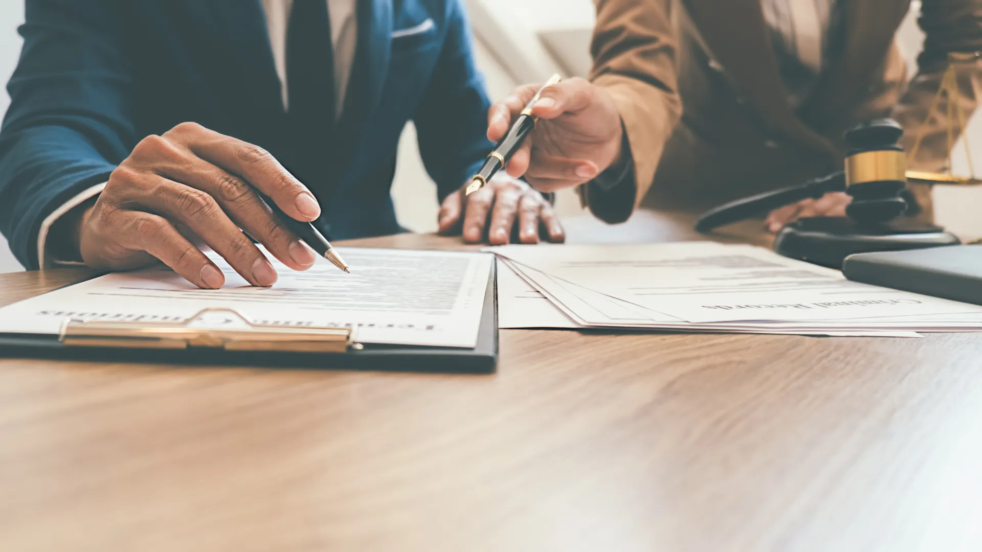 Two professionals reviewing and pointing at legal documents on a clipboard at a wooden desk.