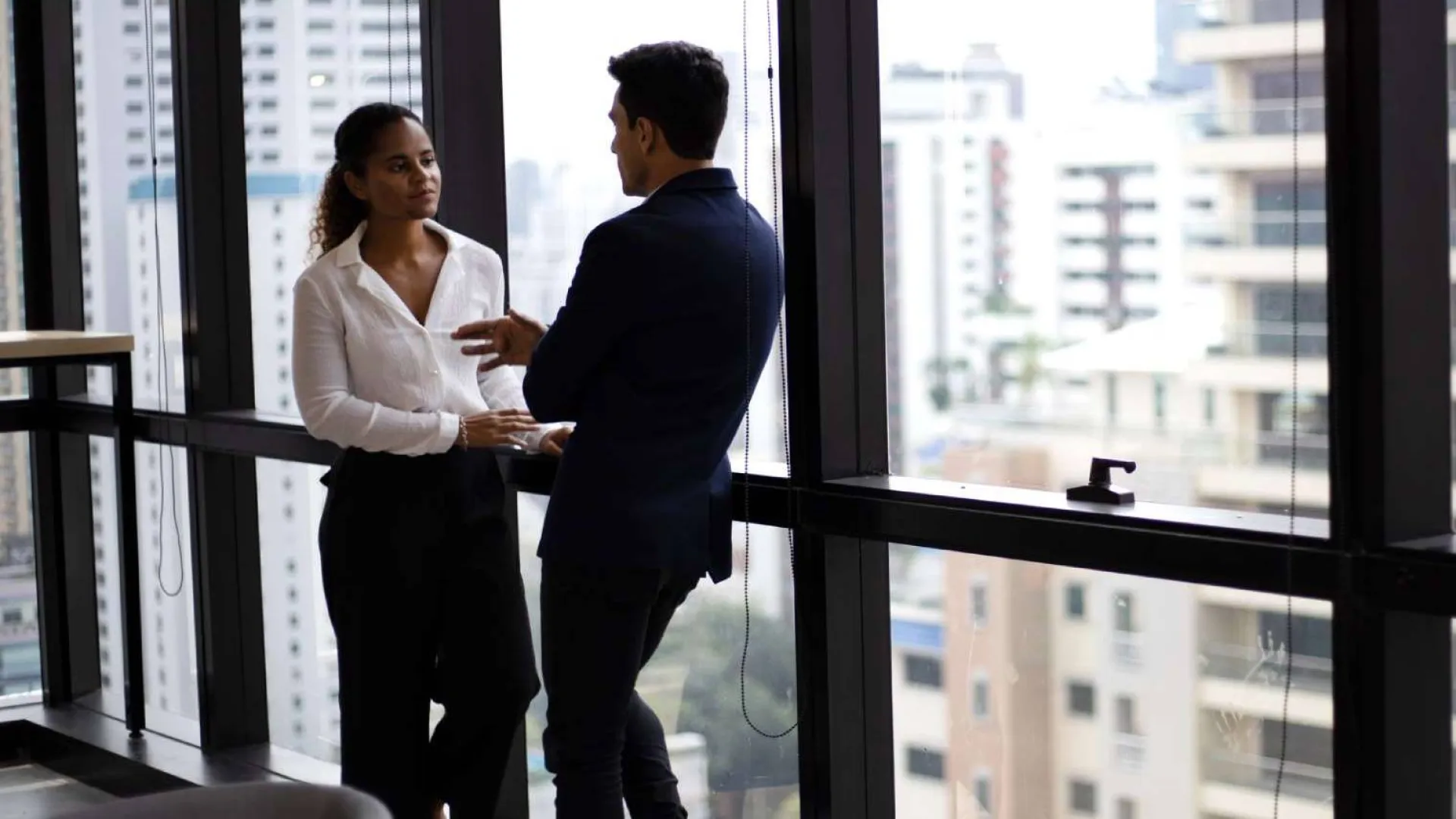 Two professionals engage in a serious discussion by a large office window overlooking the city skyline.