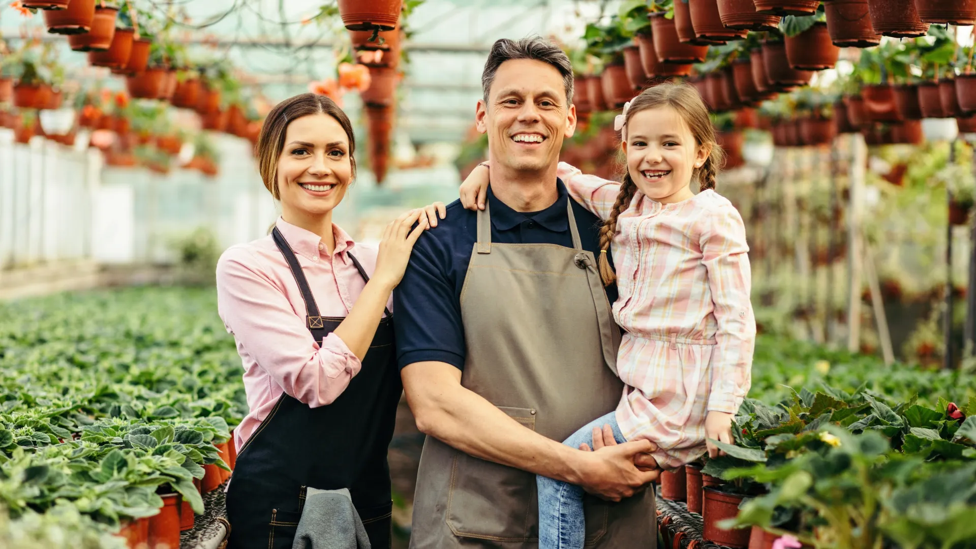 Smiling family of three in aprons standing in greenhouse surrounded by potted plants and hanging pots