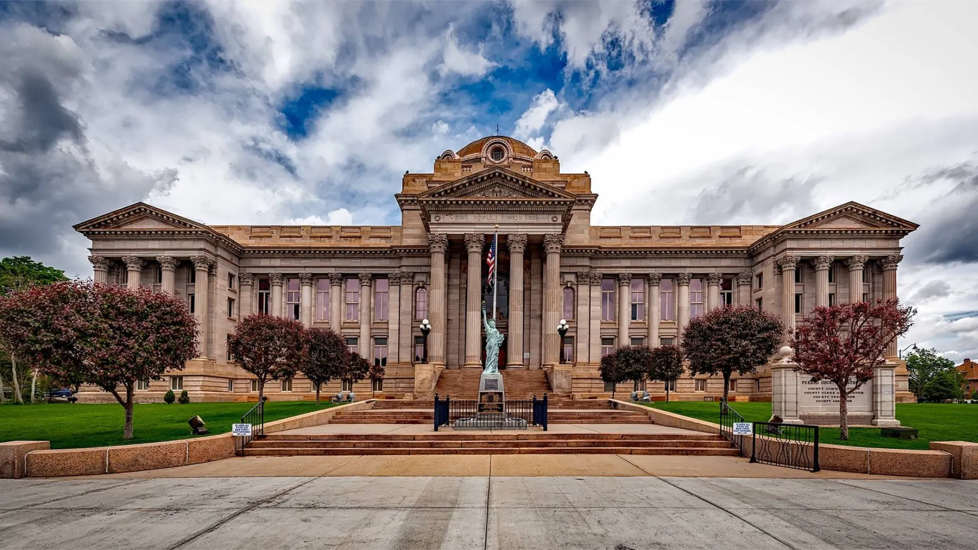 Grand neoclassical government building with columns, statue, trees, and dramatic cloudy sky overhead
