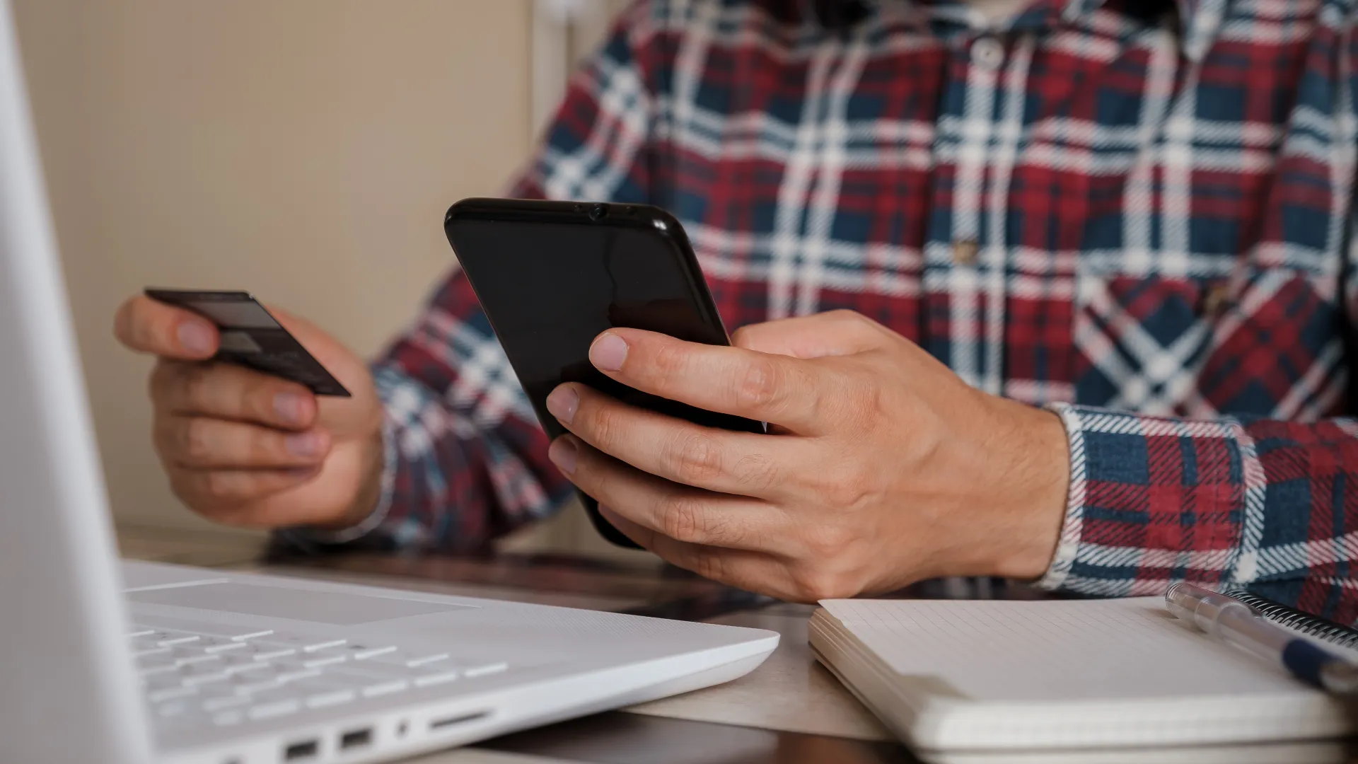 Person shopping online with a smartphone and credit card, laptop on table, casual attire highlighting modern technology.