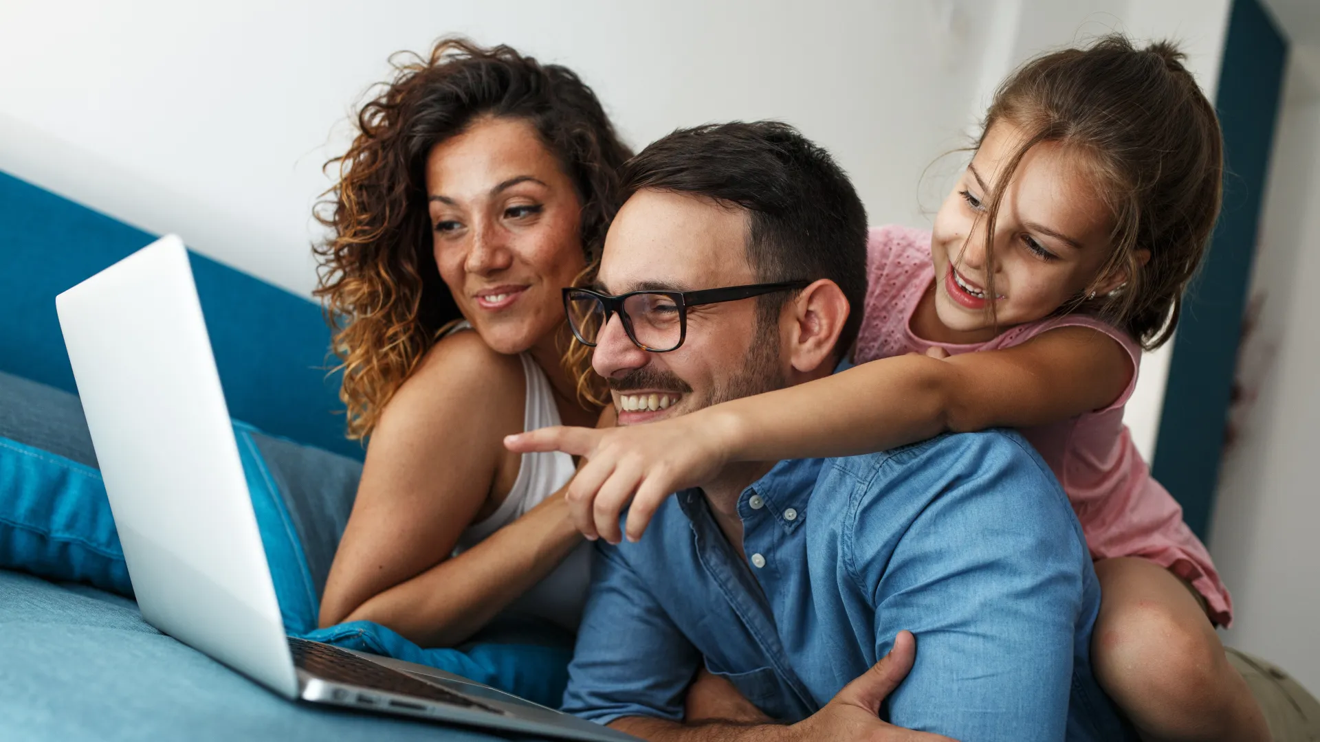 Happy family of three using a laptop together on a blue couch, smiling and pointing at the screen.