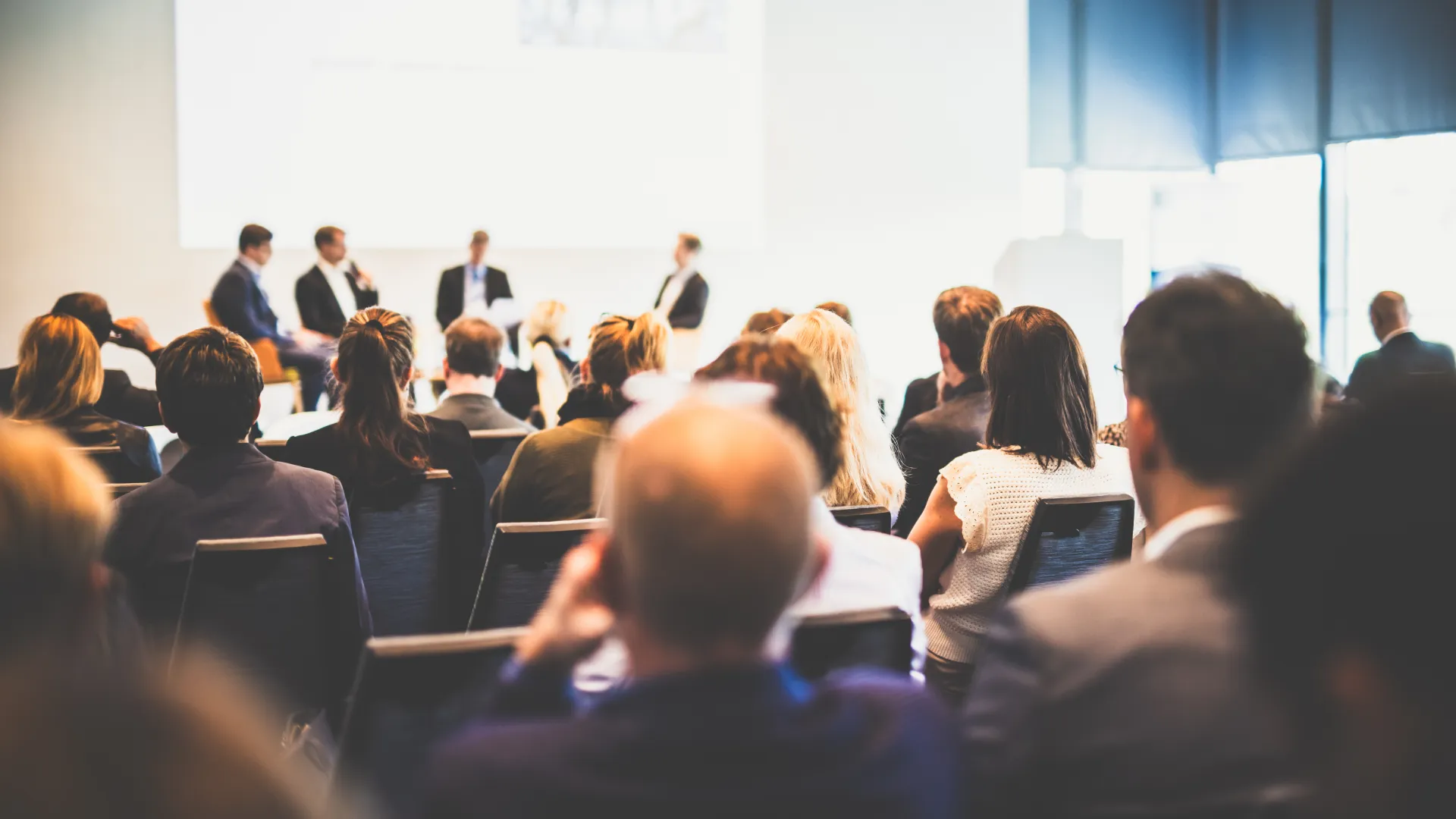 Audience attending a business conference panel discussion in a modern, bright meeting room.