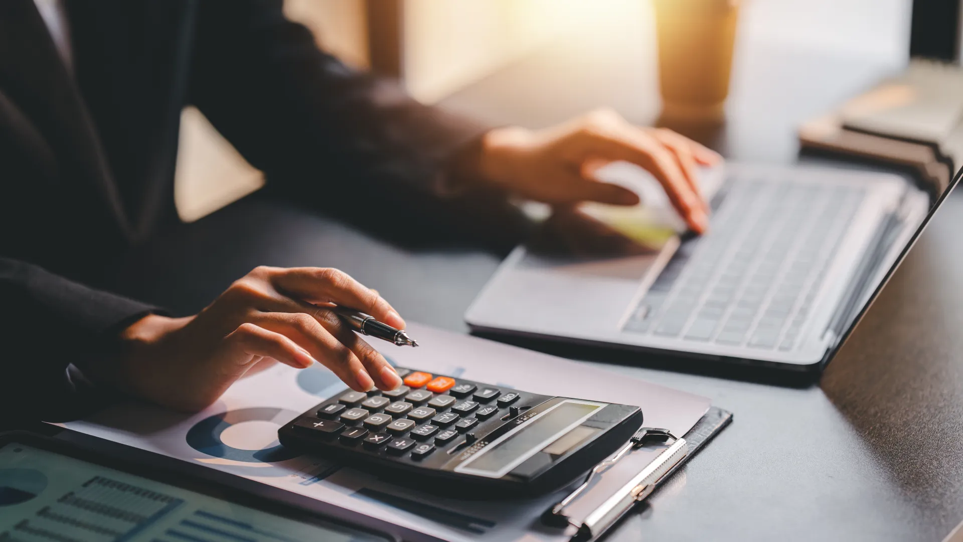 Person using calculator and laptop on desk with financial charts and documents in warm light.
