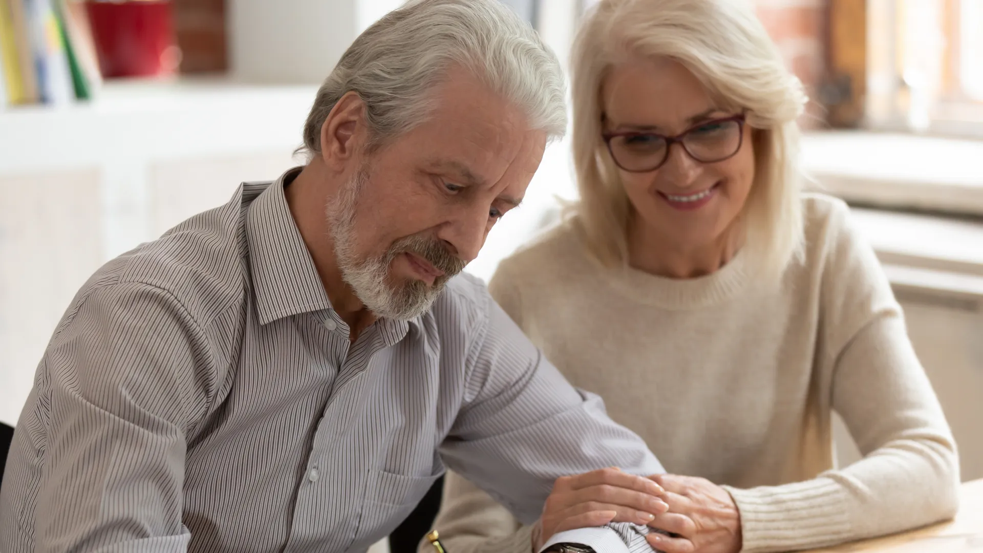 Senior man signing document while woman supports him, seated at table in cozy indoor setting.