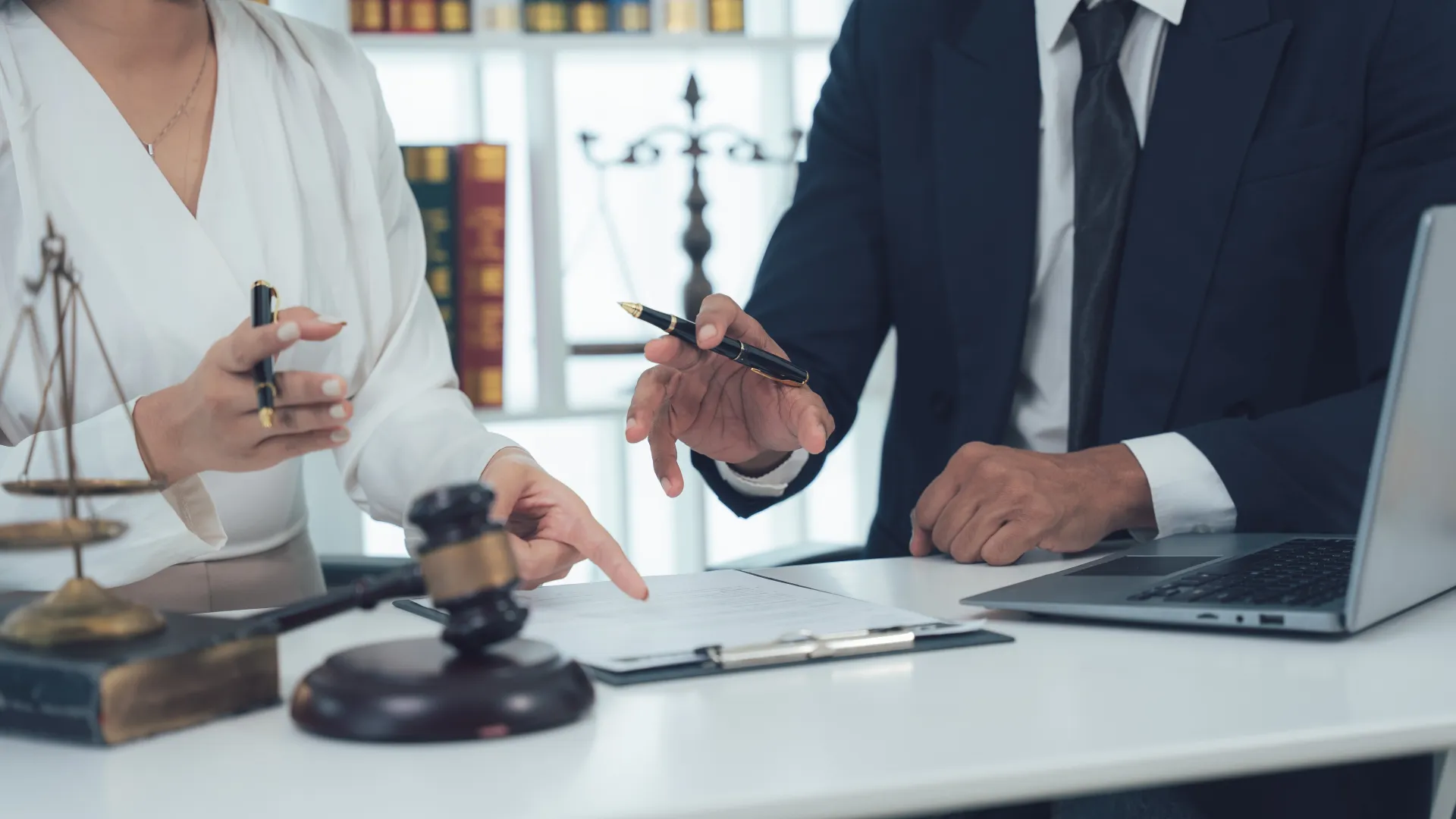 Two lawyers discussing documents at a desk with a gavel, legal books, and a laptop in a law office.