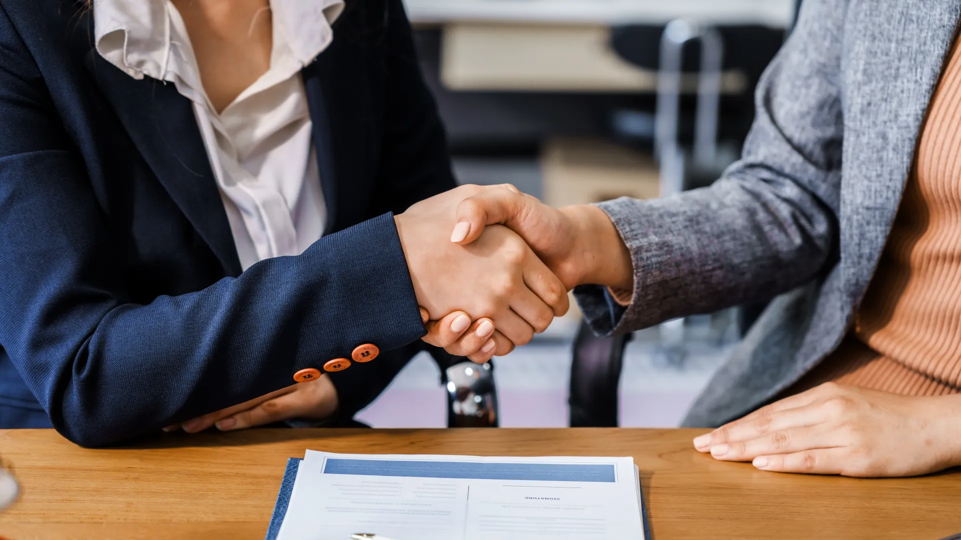 Two businesswomen shaking hands over a desk with contract papers and pen, symbolizing agreement and partnership.