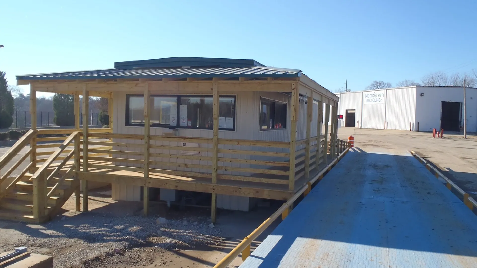 Small wooden office building with a porch beside a large blue vehicle scale in an industrial yard under clear skies