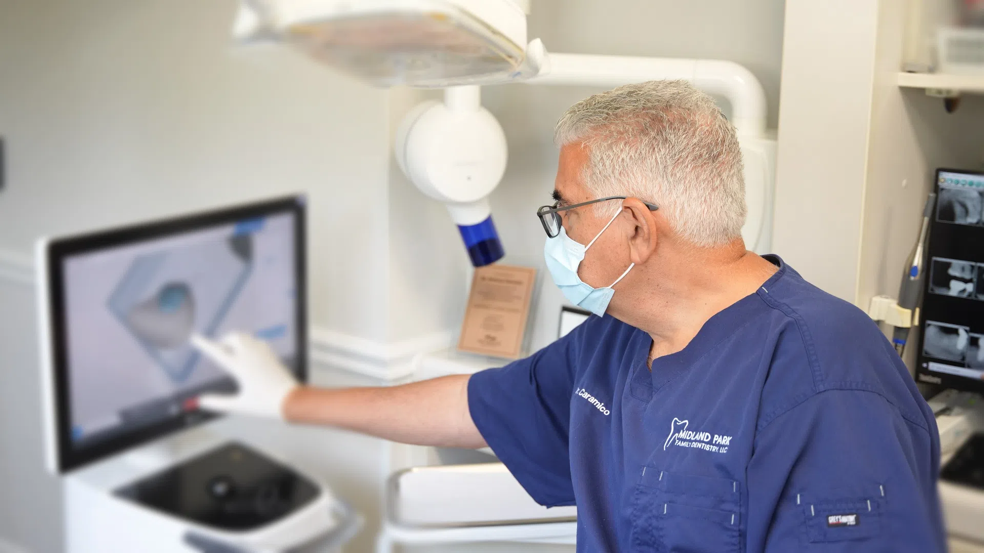 Dentist in blue scrubs using digital imaging technology to examine a 3D dental model on a screen.
