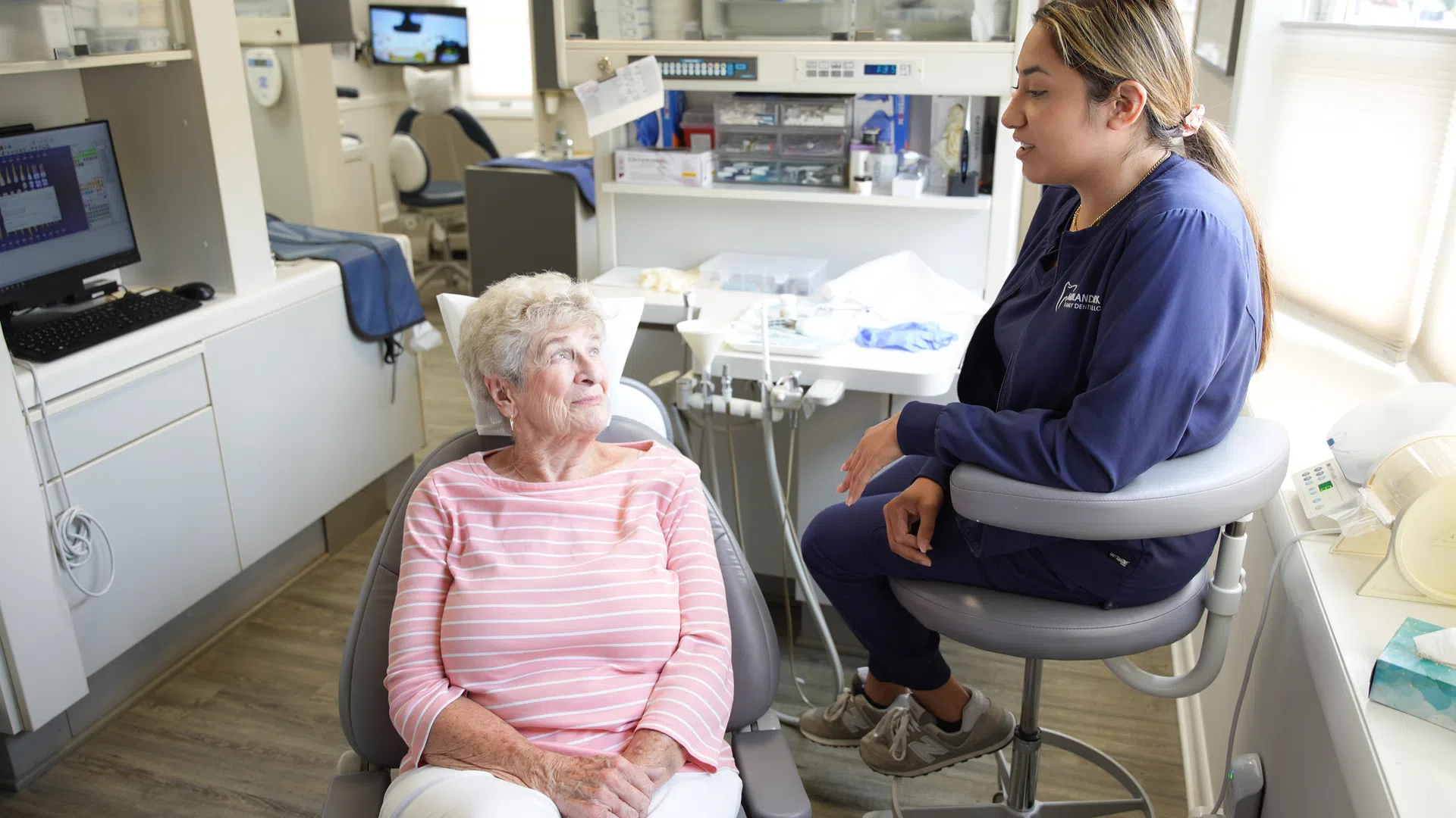 Elderly woman in pink striped shirt sitting and talking with a female dental professional in a dental office.