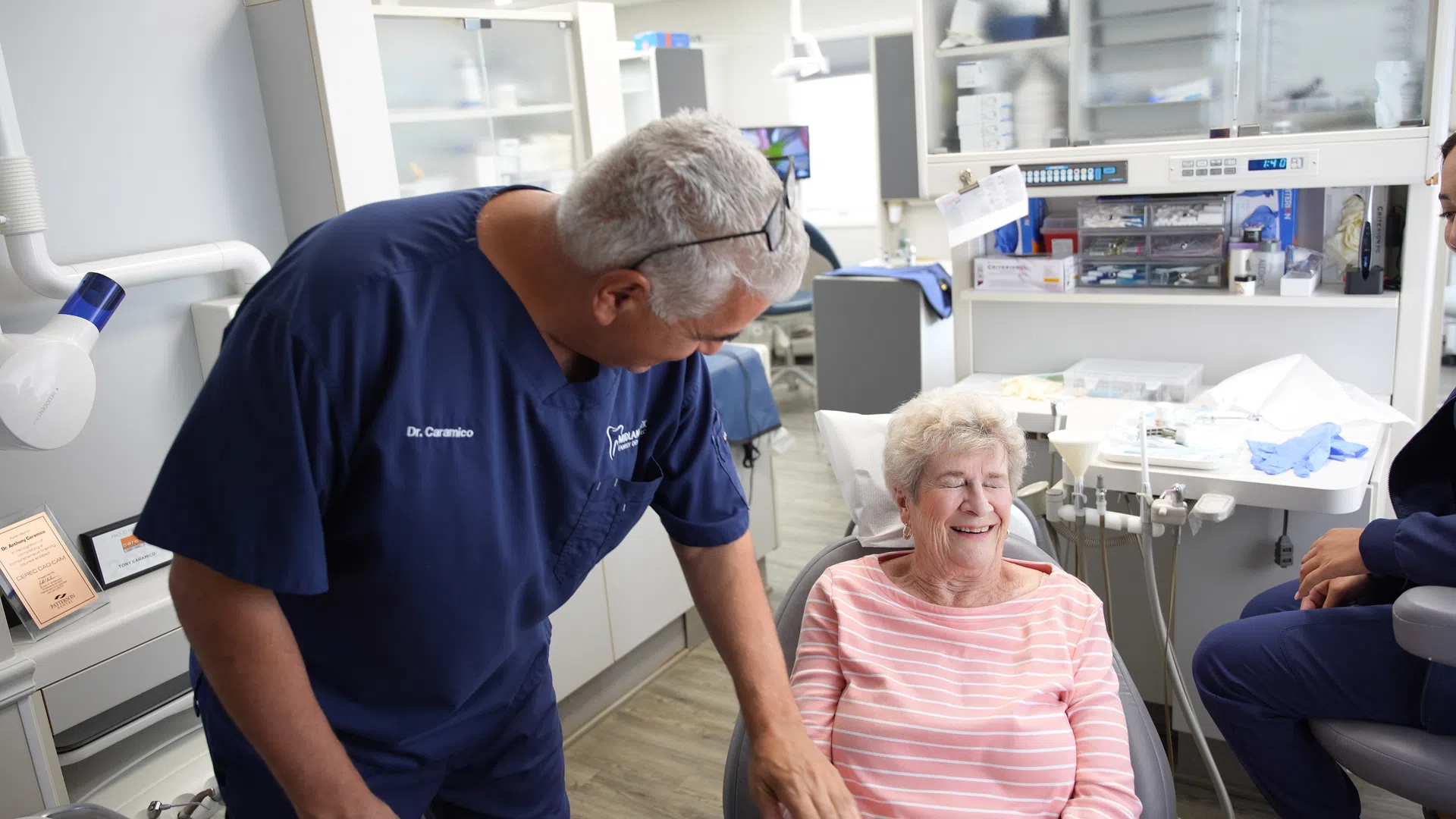 Dentist in navy scrubs smiling and talking to elderly female patient in dental chair at clinic.